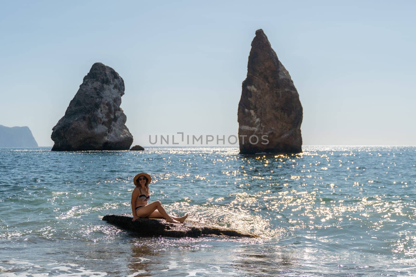 A woman sits on a rock in the ocean, facing two large rock formations in the distance. The water is blue and the sky is clear.