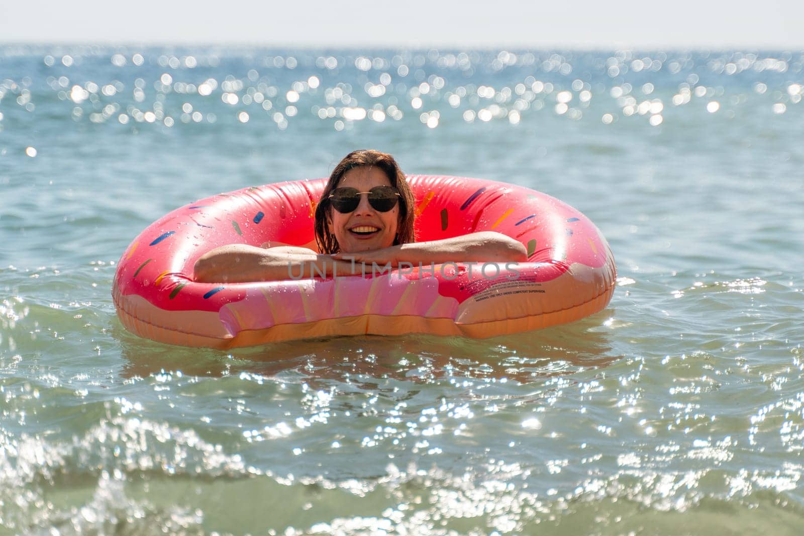 A woman floats in the ocean on a pink inflatable ring. She is wearing sunglasses and smiling at the camera. The water is blue and sparkling.