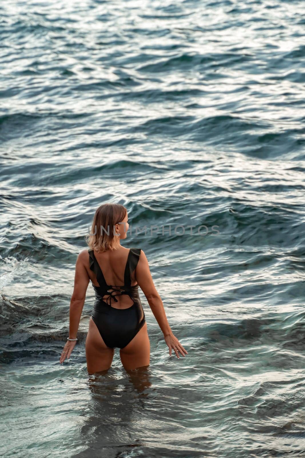 A woman in a black swimsuit stands in the ocean, facing away from the camera, with waves splashing around her.