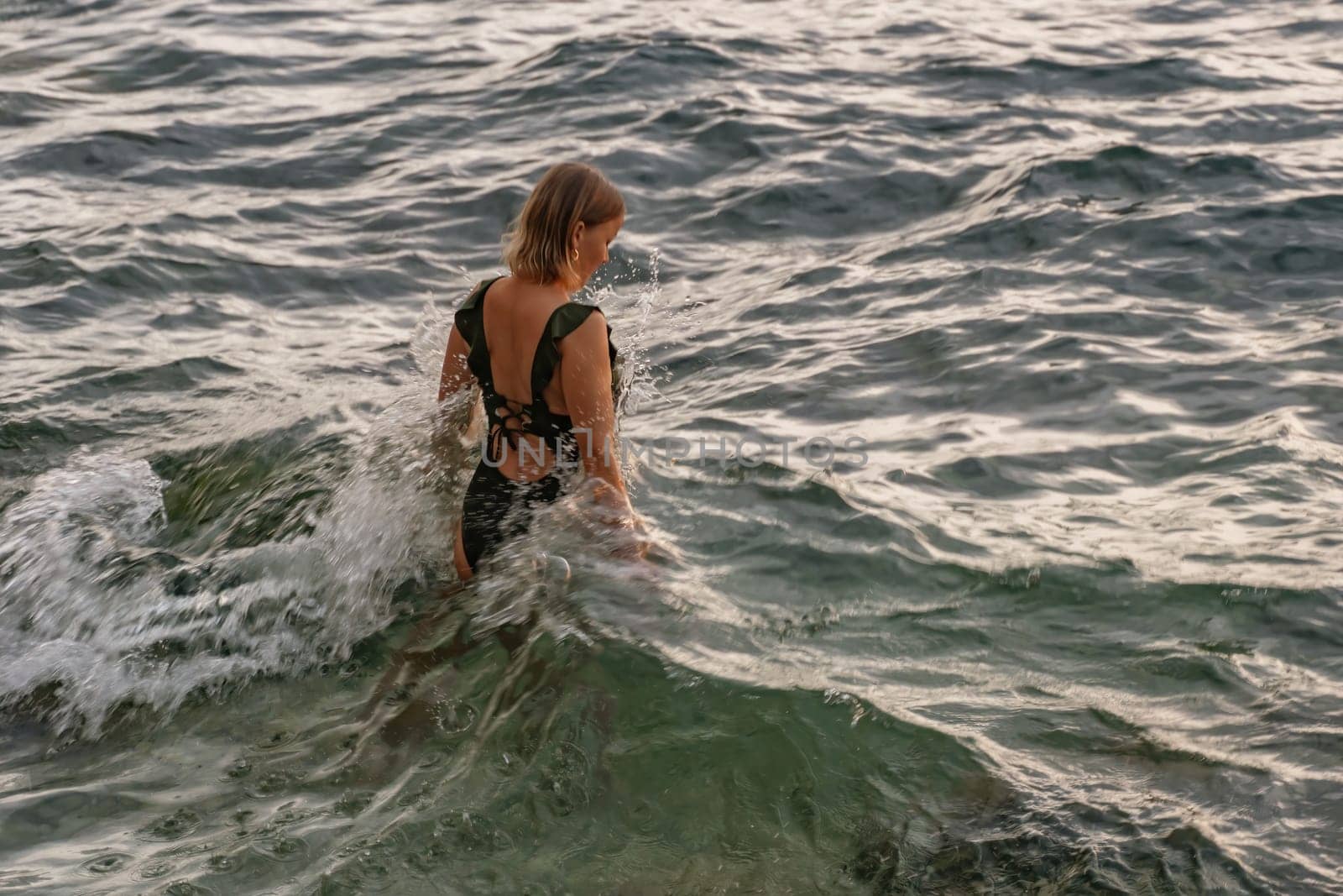 A woman in a black swimsuit walks through choppy waves, her back to the camera. The water is splashing around her.