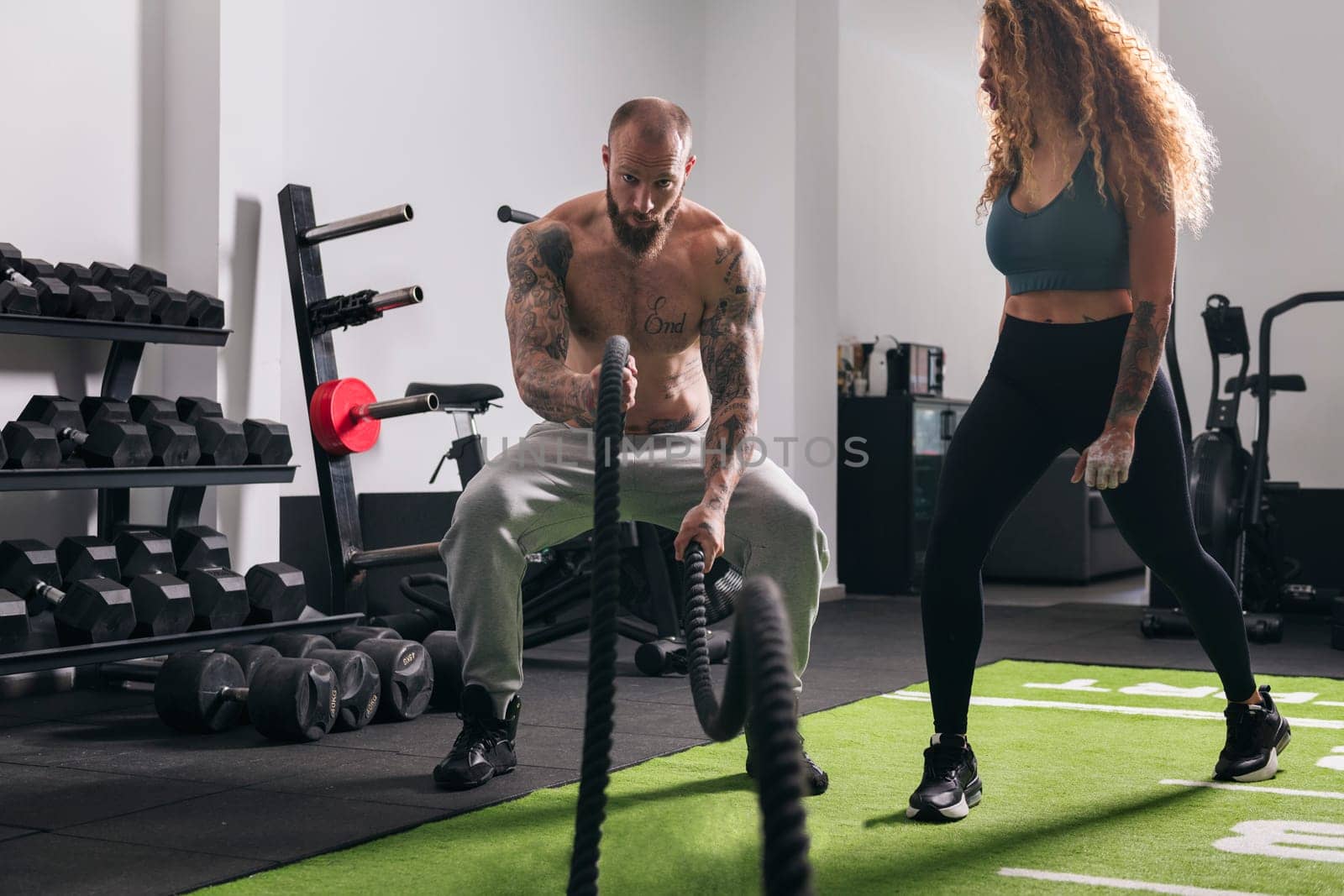muscular sportsman with tattoos doing exercise with rope during functional workout with female instructor at gym, concept of fitness and active lifestyle