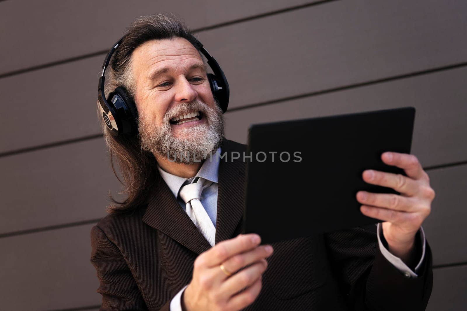 content mature male with beard in formal suit and headphones using tablet while laughing communicating online against gray wall, concept of middle age business man and technology of communication