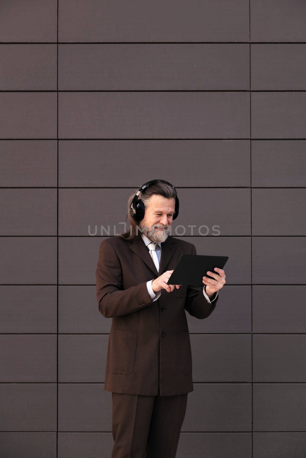 content mature male with beard in formal suit and wireless headphones using tablet while communicating online against gray wall, concept of middle age business man and technology of communication