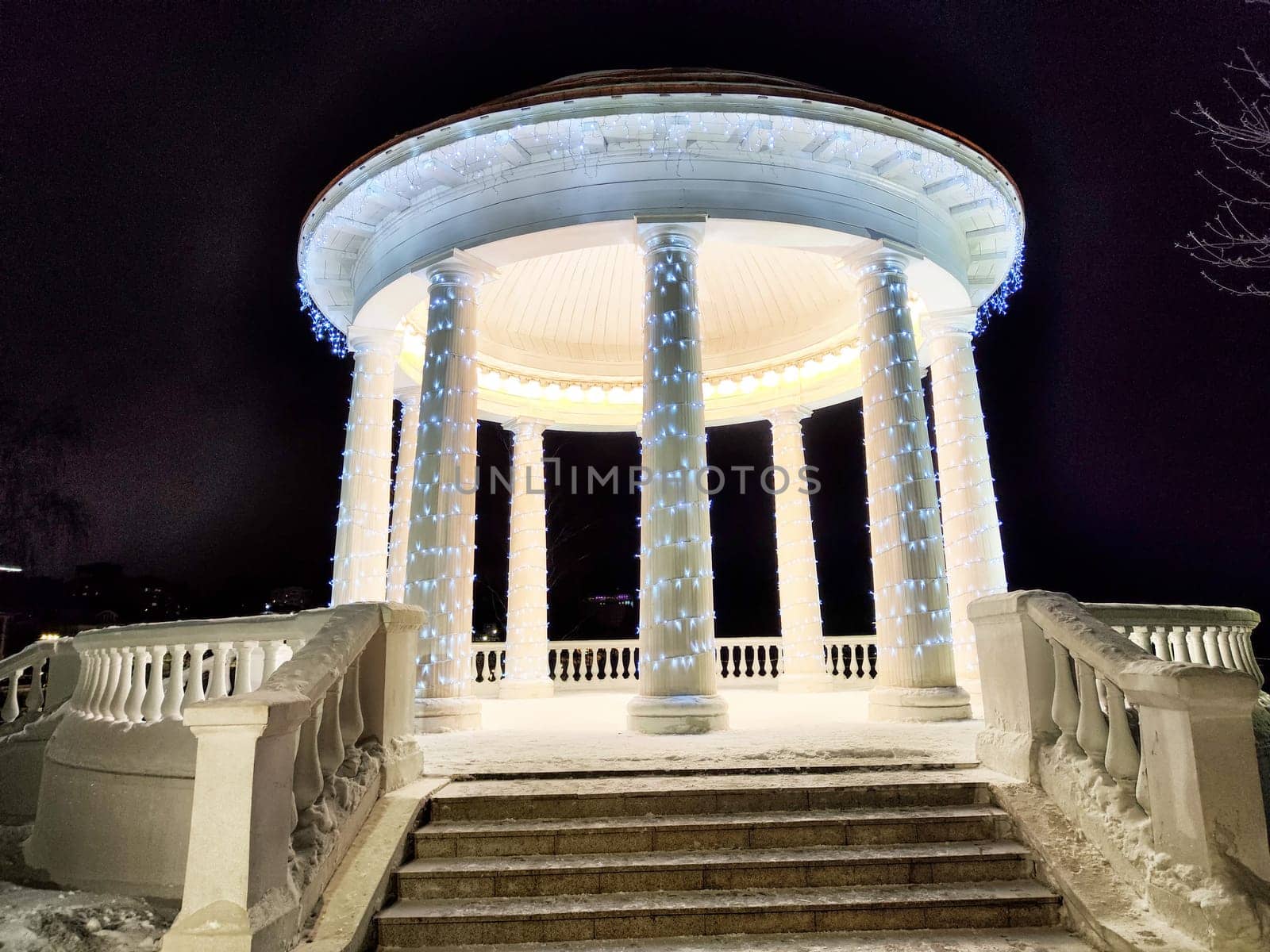 Illuminated gazebo with columns shines brightly in a snowy park during winter night, creating a serene and magical atmosphere.