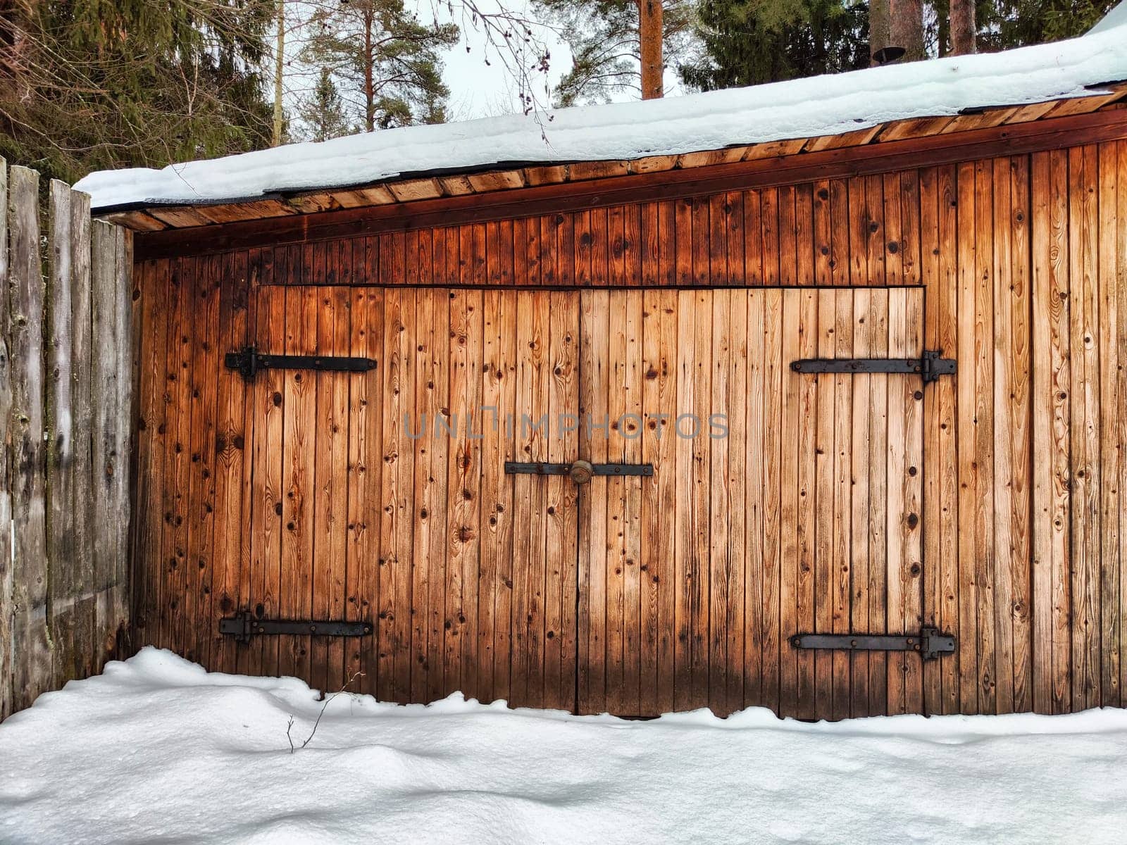 A rustic wooden shed stands against a backdrop of snow-covered ground and pine trees, showcasing sturdy iron door fittings.