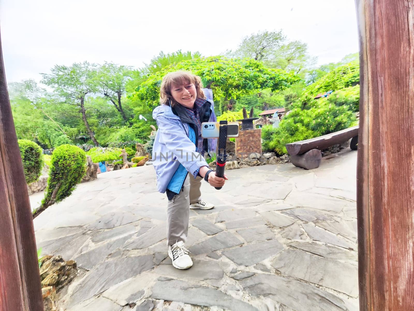 Middle-aged woman happily takes a selfie in a curved mirror while exploring a beautiful park filled with nature.