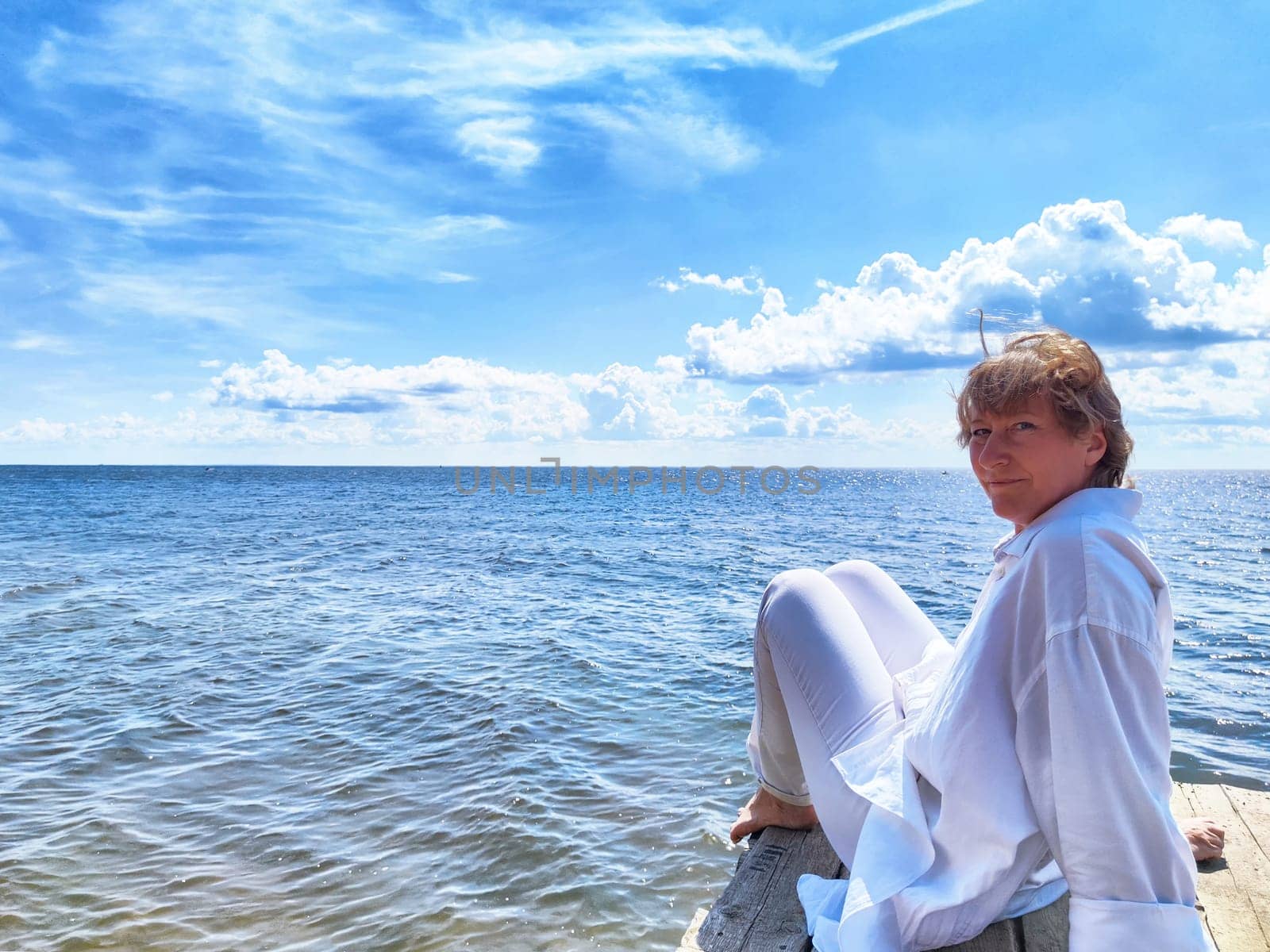 Blonde woman relaxes on a dock, gazing thoughtfully at the expansive ocean while embracing the peacefulness of nature.
