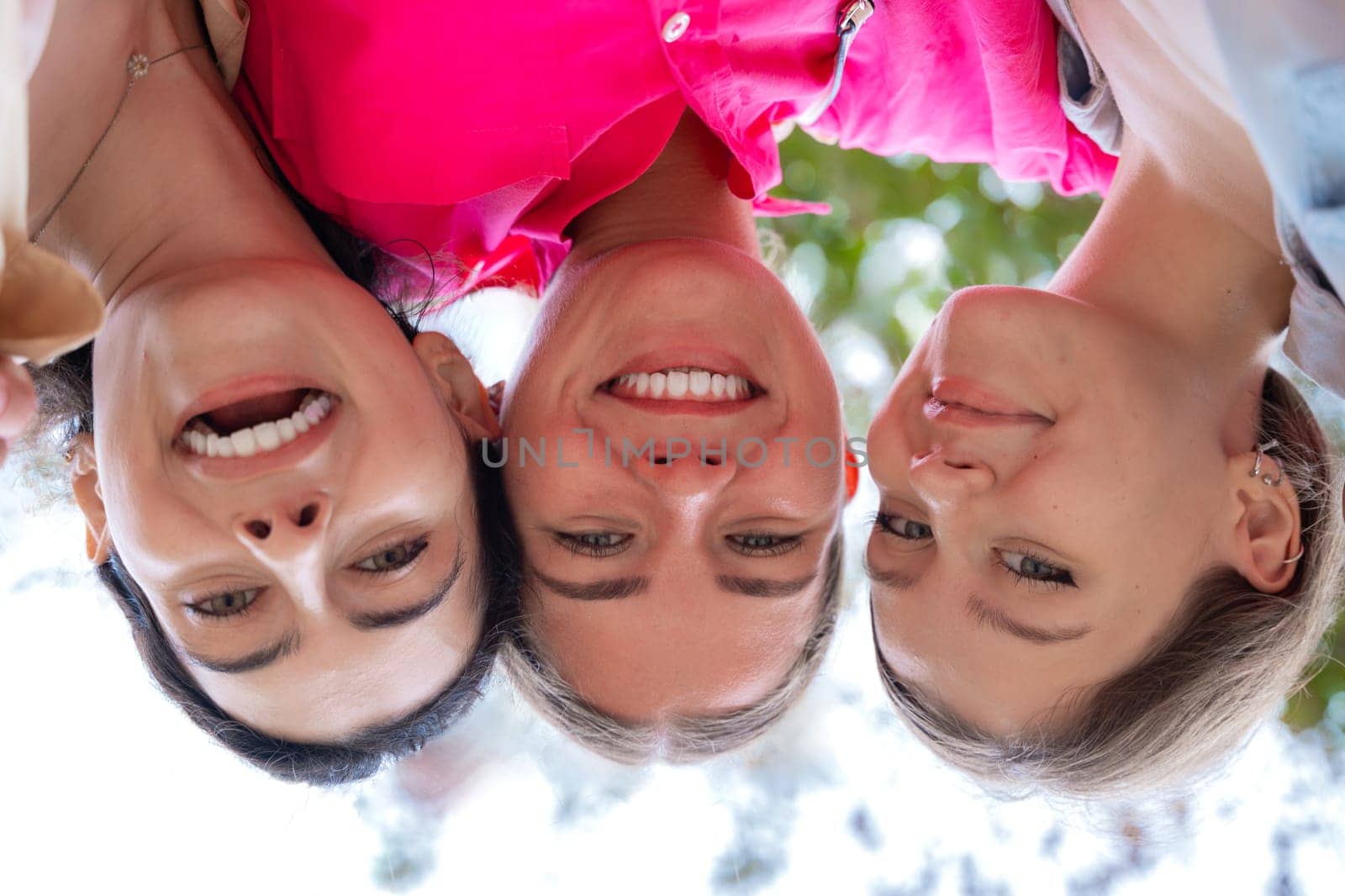 Three young women laughing and smiling while posing upside down for a fun and creative photo. They appear joyful and playful. Natural outdoor background.
