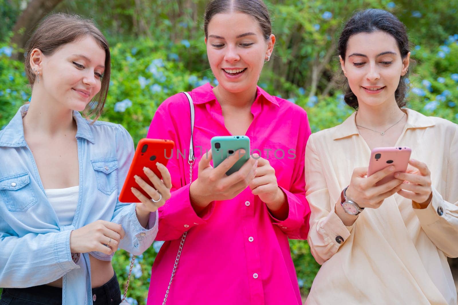 Three young women standing outdoors, smiling while looking at their smartphones. They appear to be enjoying a conversation or sharing content. Casual clothing and green background.