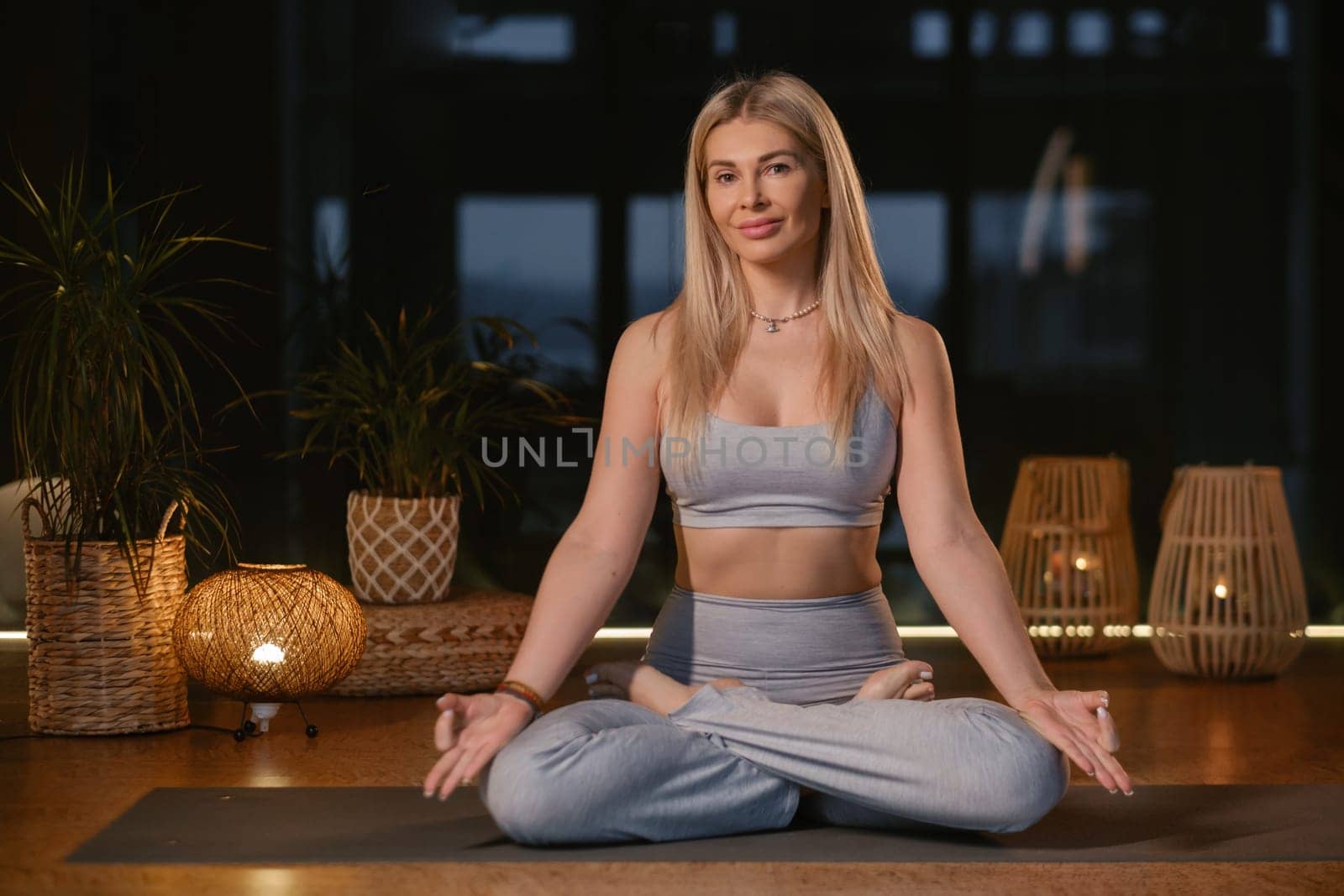 A woman is engaged in a yoga practice within a contemporary studio, surrounded by ambient lighting and decorative elements. She sits cross-legged on a mat, hands positioned in prayer, embodying tranquility and focus.