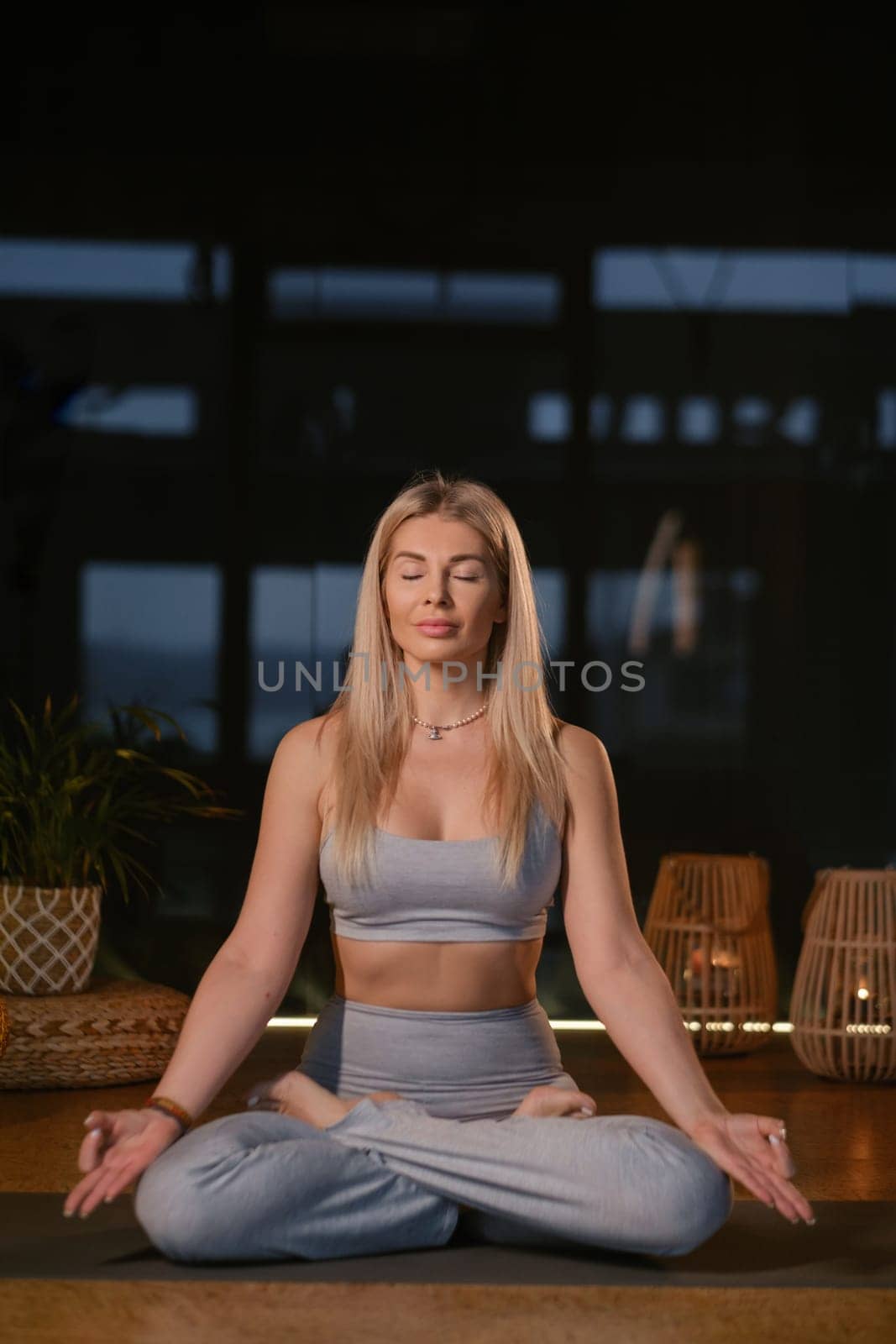 A woman sits cross-legged on a mat, eyes closed in deep concentration, in a tranquil indoor environment. Soft lighting and natural decor create a peaceful atmosphere, ideal for meditation during the evening.