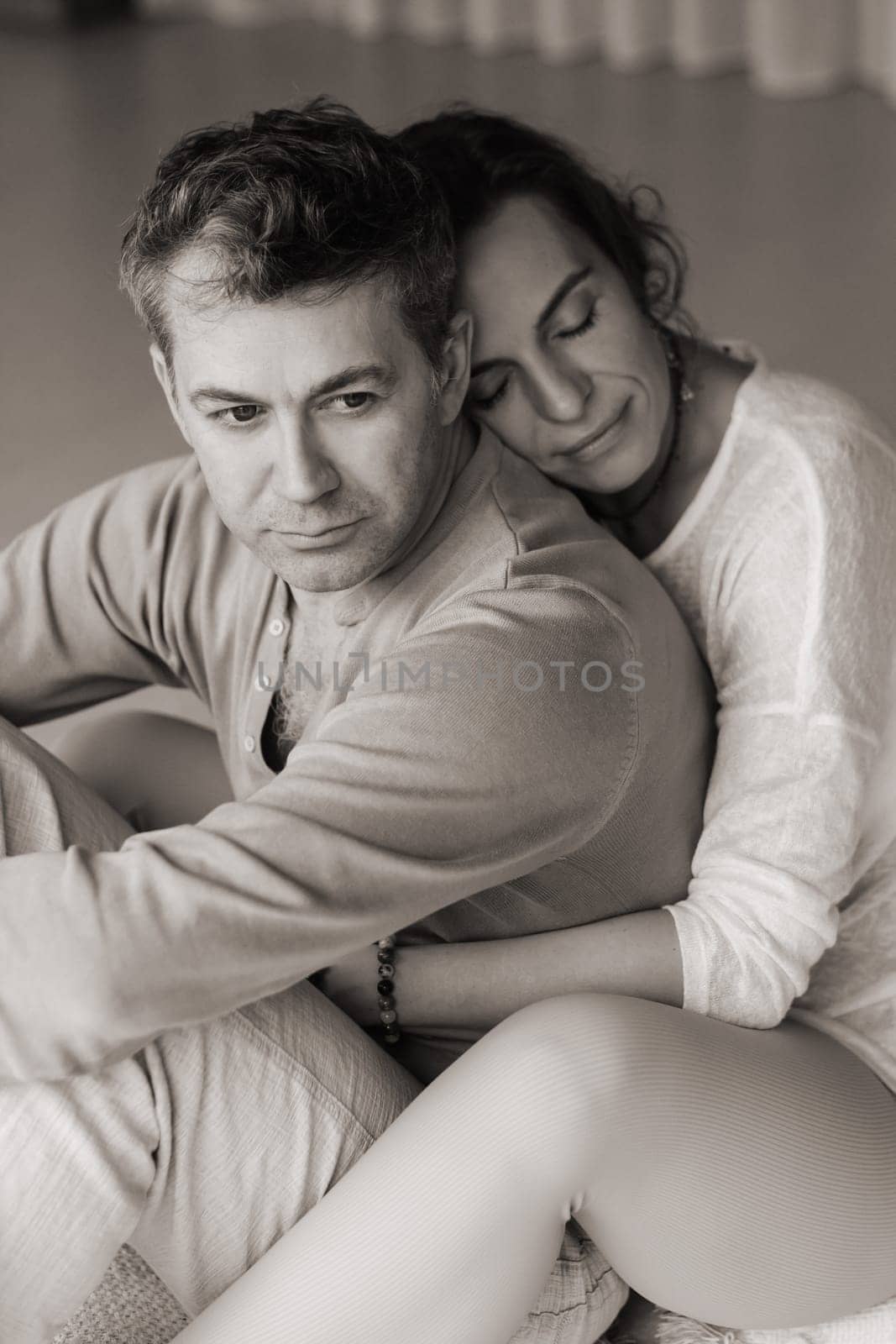 A man and woman sit back to back on a soft mat, surrounded by a tranquil indoor environment. The warm afternoon sunlight filters through large windows, creating a peaceful atmosphere as they focus on their mindfulness meditation practice together.