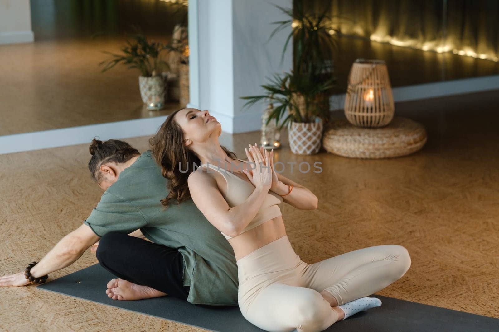 A couple engages in partner yoga in a tranquil indoor studio. They sit back-to-back, demonstrating a pose that emphasizes balance and trust between each other. Soft lighting adds to the calming ambiance of the space.