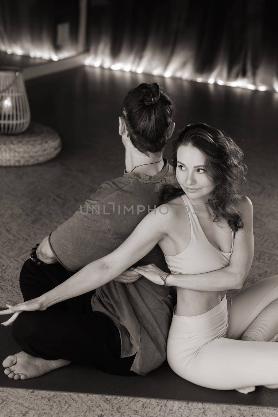 A couple engages in partner yoga in a tranquil indoor studio. They sit back-to-back, demonstrating a pose that emphasizes balance and trust between each other. Soft lighting adds to the calming ambiance of the space.