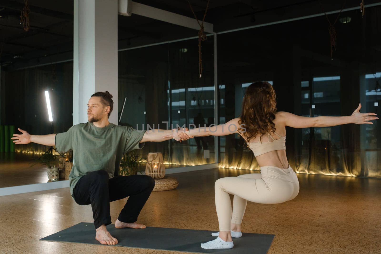 A couple engages in partner yoga poses in a well-lit and spacious studio. They hold hands while balancing on one leg, demonstrating teamwork and coordination. Natural light streams through large windows, enhancing the serene atmosphere.