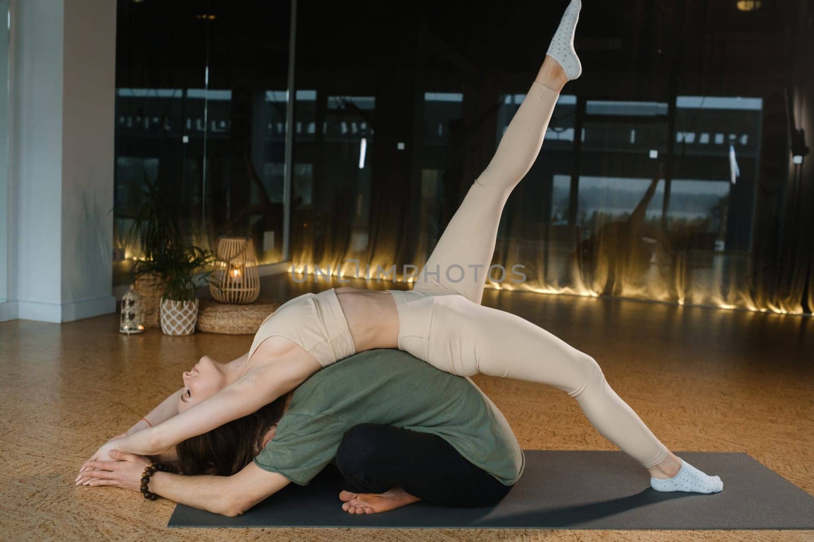 A woman balances gracefully in a difficult yoga pose while resting on the back of a man, who supports her with stability. The modern studio features large windows and soft lighting, creating a tranquil morning atmosphere ideal for yoga.