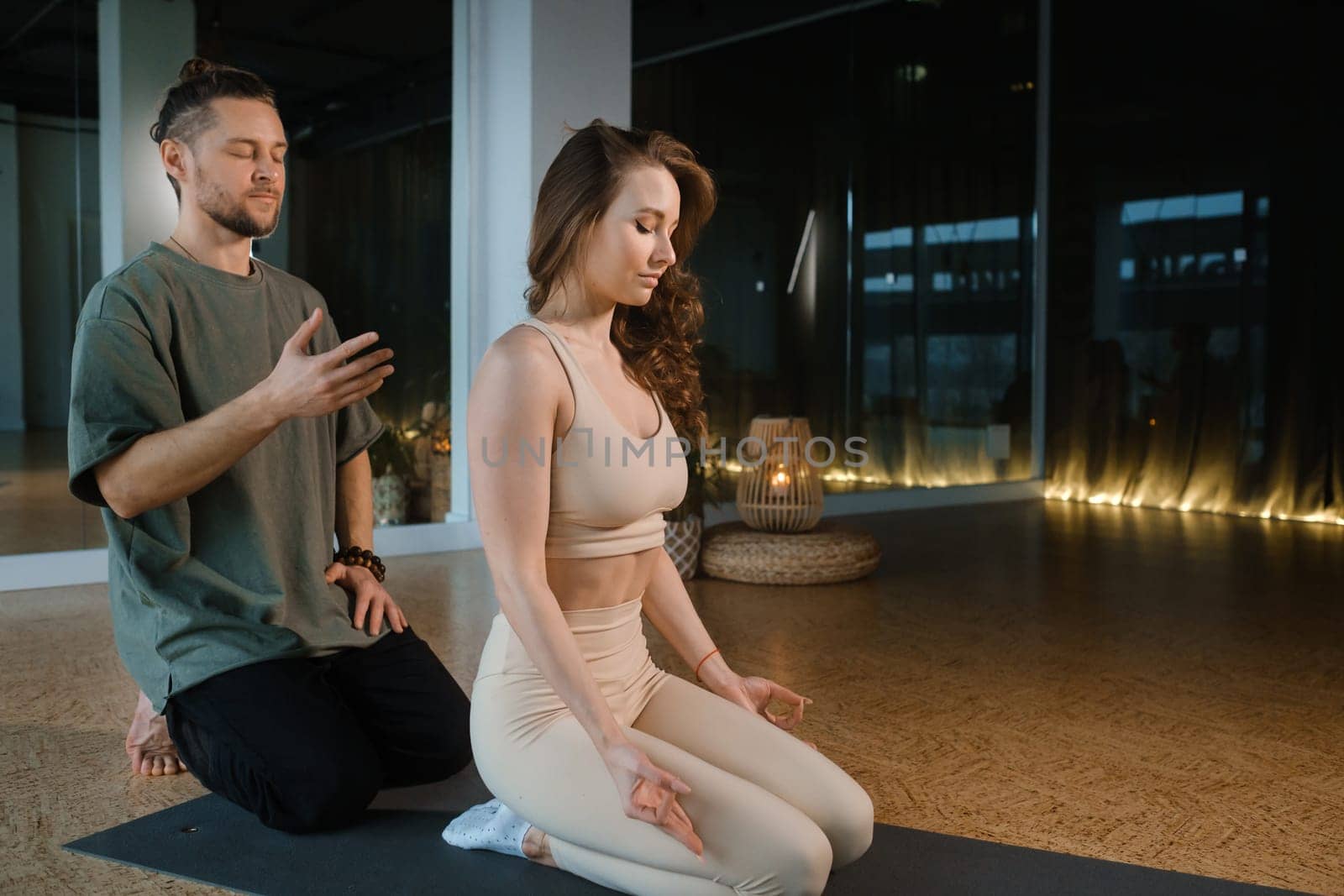 Two individuals engage in a calming yoga and meditation practice inside a serene studio. The dimly lit space features soft lighting and a peaceful atmosphere, enhancing their focus and mindfulness.