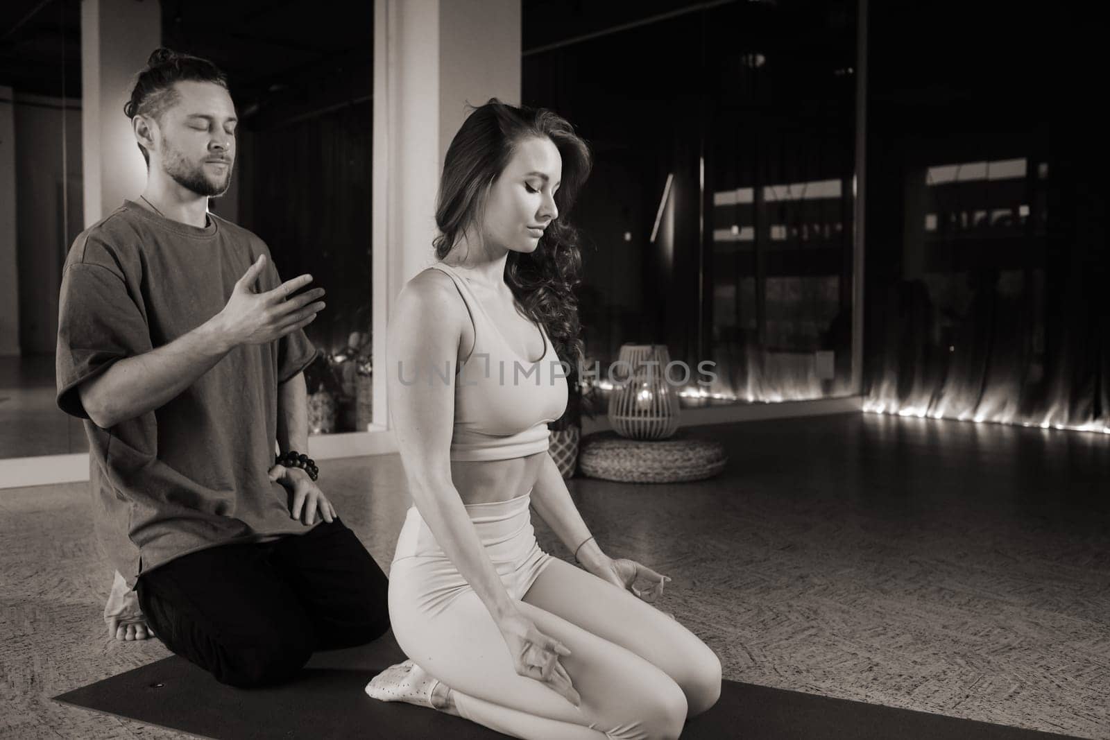 Two individuals engage in a calming yoga and meditation practice inside a serene studio. The dimly lit space features soft lighting and a peaceful atmosphere, enhancing their focus and mindfulness.