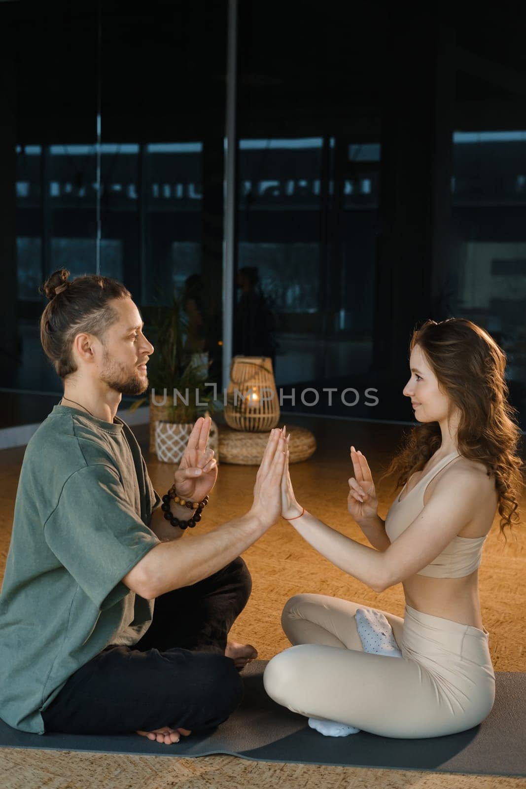 A couple sits cross-legged on mats in a serene yoga studio, gently pressing their palms together in a synchronized motion. The warm light highlights their calm expressions as they engage in a mindful practice, fostering a deep sense of connection.