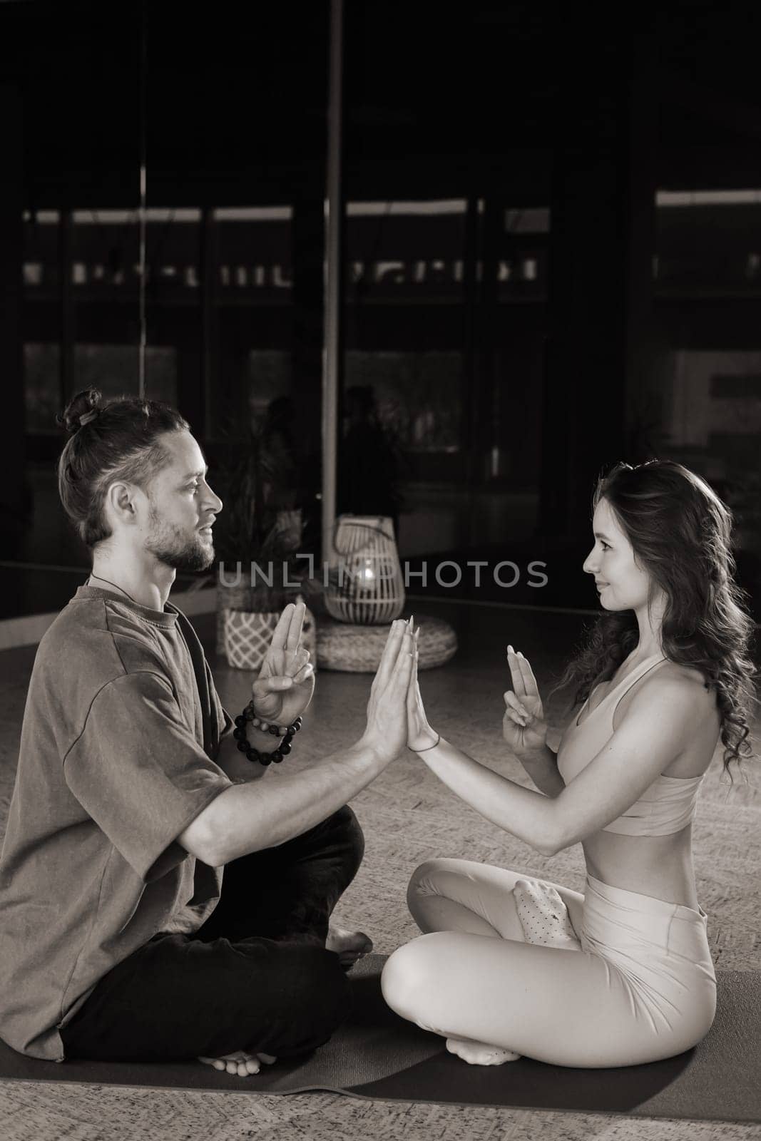 A couple sits cross-legged on mats in a serene yoga studio, gently pressing their palms together in a synchronized motion. The warm light highlights their calm expressions as they engage in a mindful practice, fostering a deep sense of connection.
