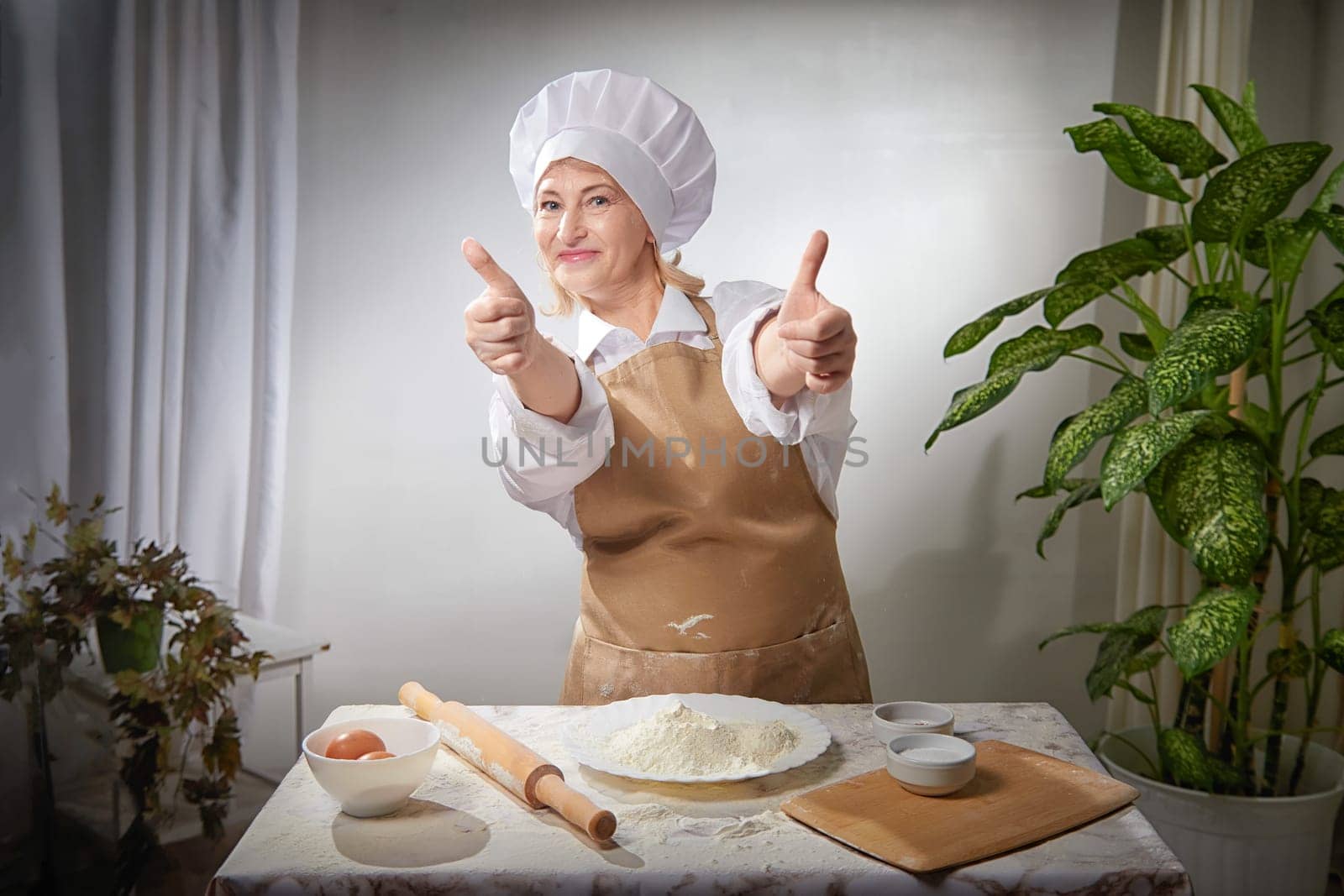 An elderly woman expertly prepares flour in a warm kitchen while playfully posing with a big smile and thumbs up.