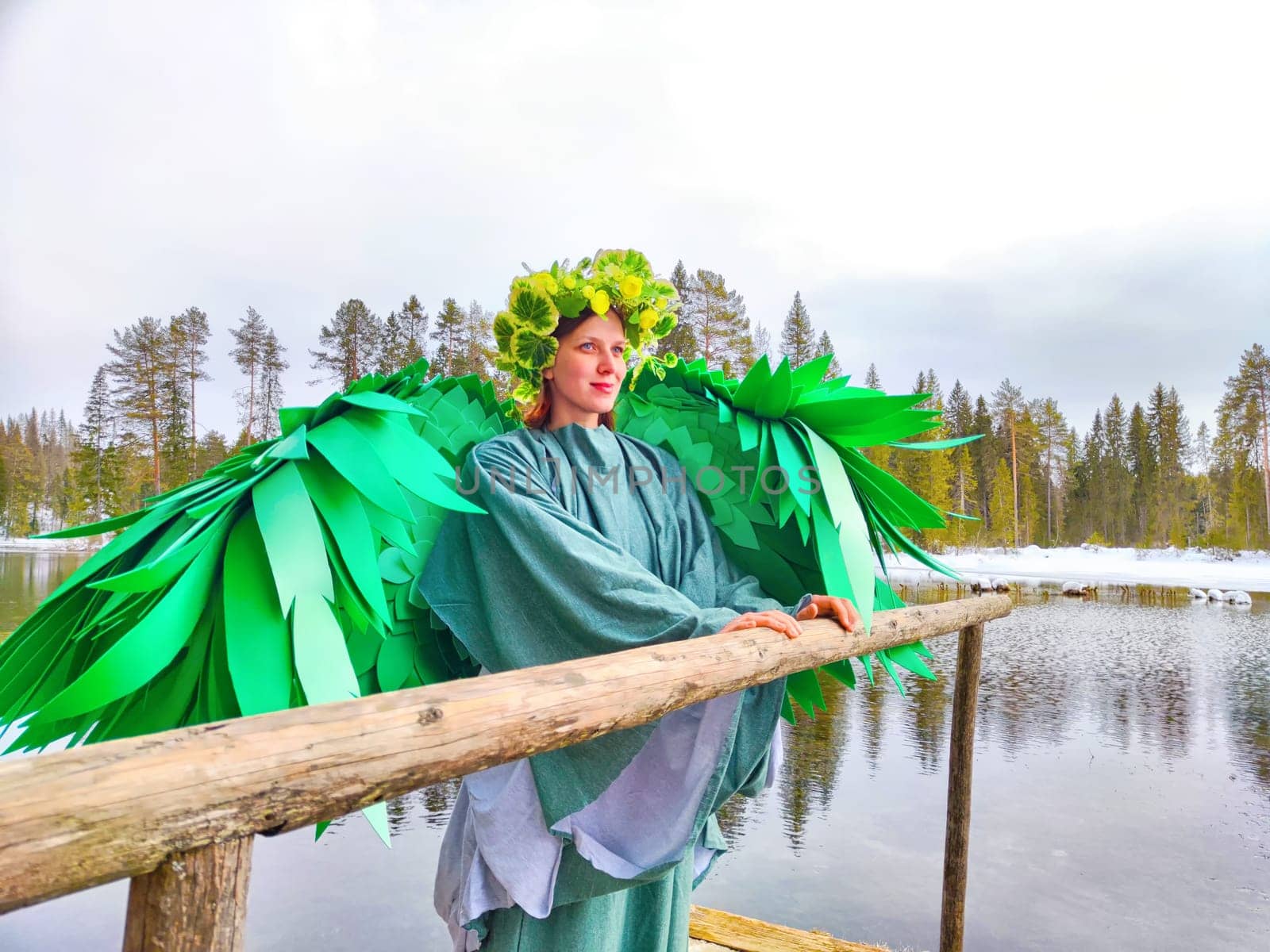 A girl in a green dress poses at a tranquil lake, embodying spring's beauty with angel-like wings, celebrating nature's renewal.
