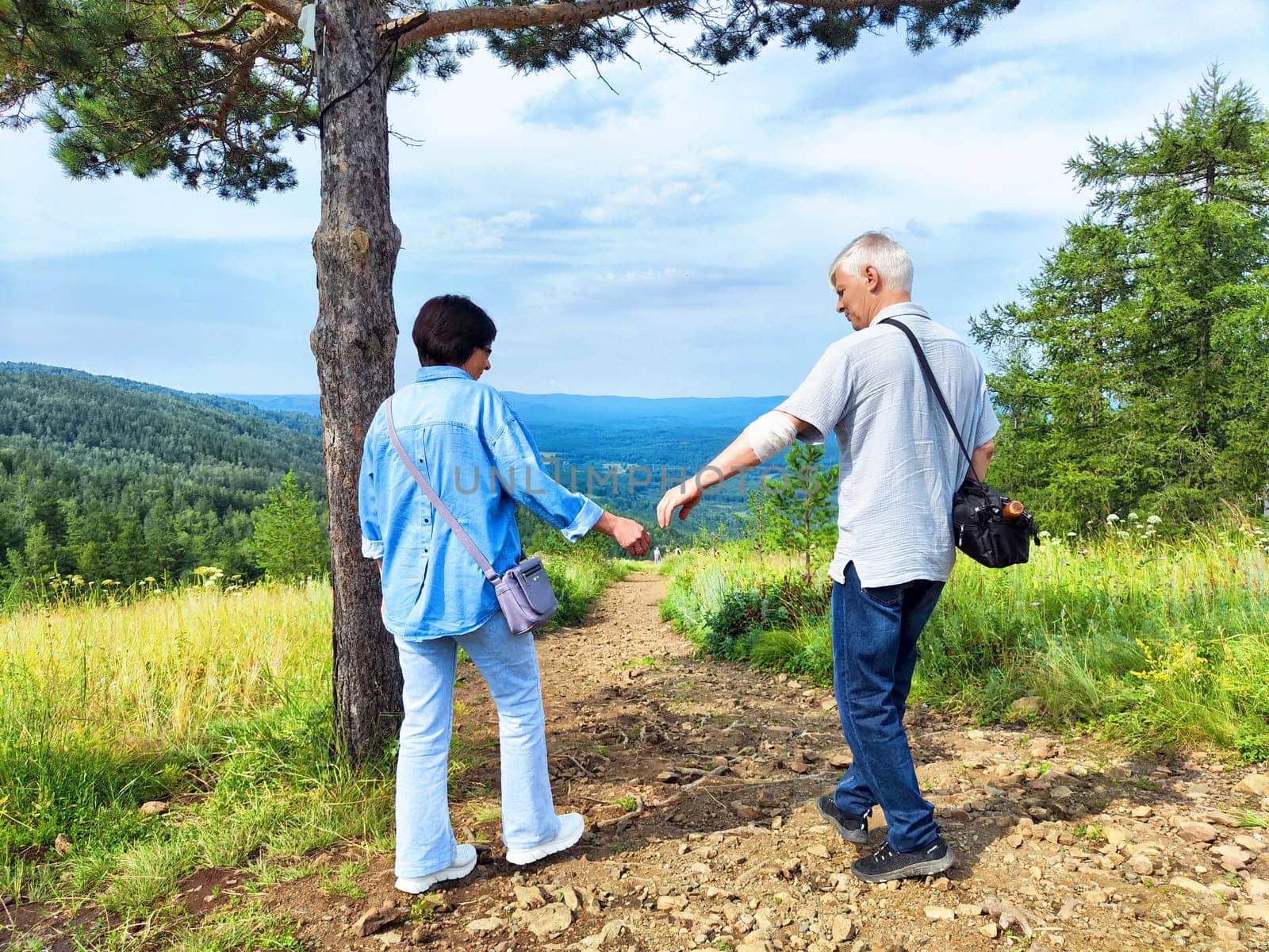 Two people stroll along a scenic trail, enjoying the view of lush hills and a blue sky on a sunny day.