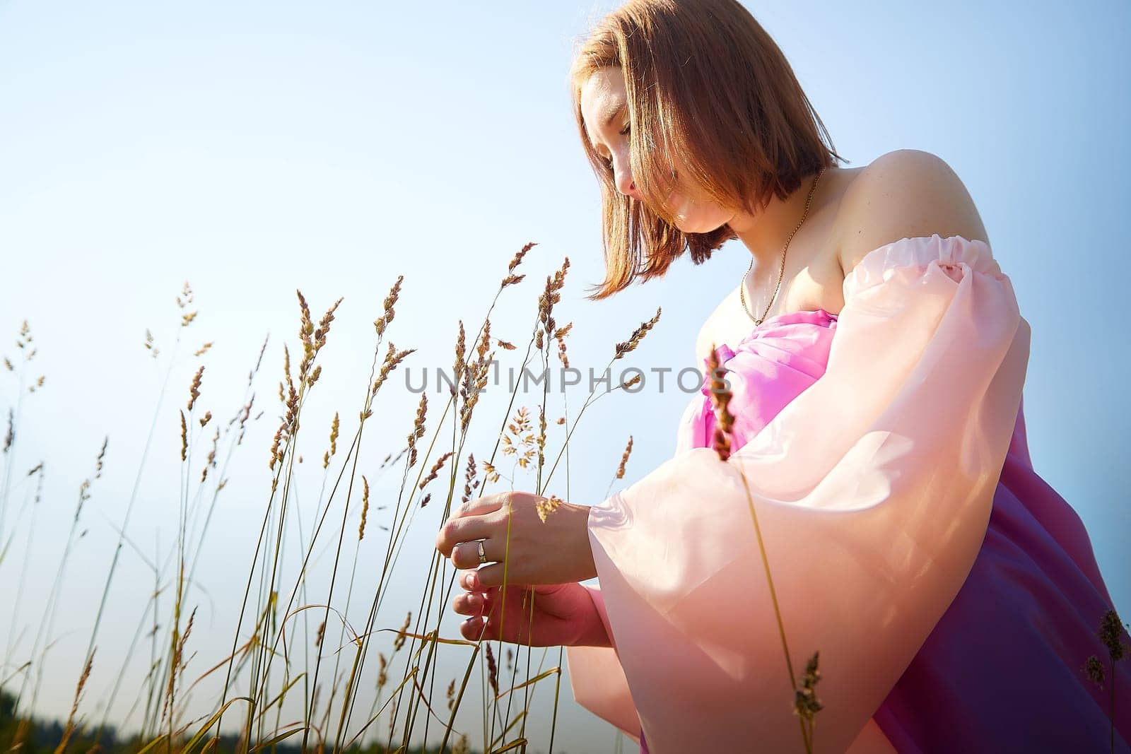 A woman in a colorful dress bends down to gather grass in a sunny field, surrounded by tall plants and clear blue skies.