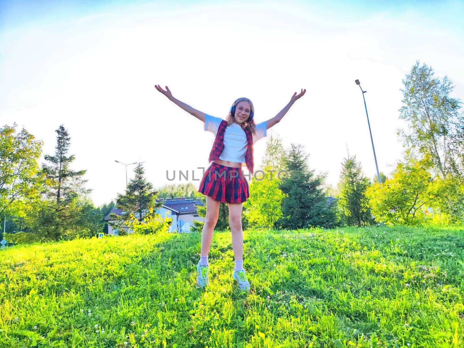 A cheerful girl stands on a grassy hill, arms raised with excitement, surrounded by trees and a bright blue sky.