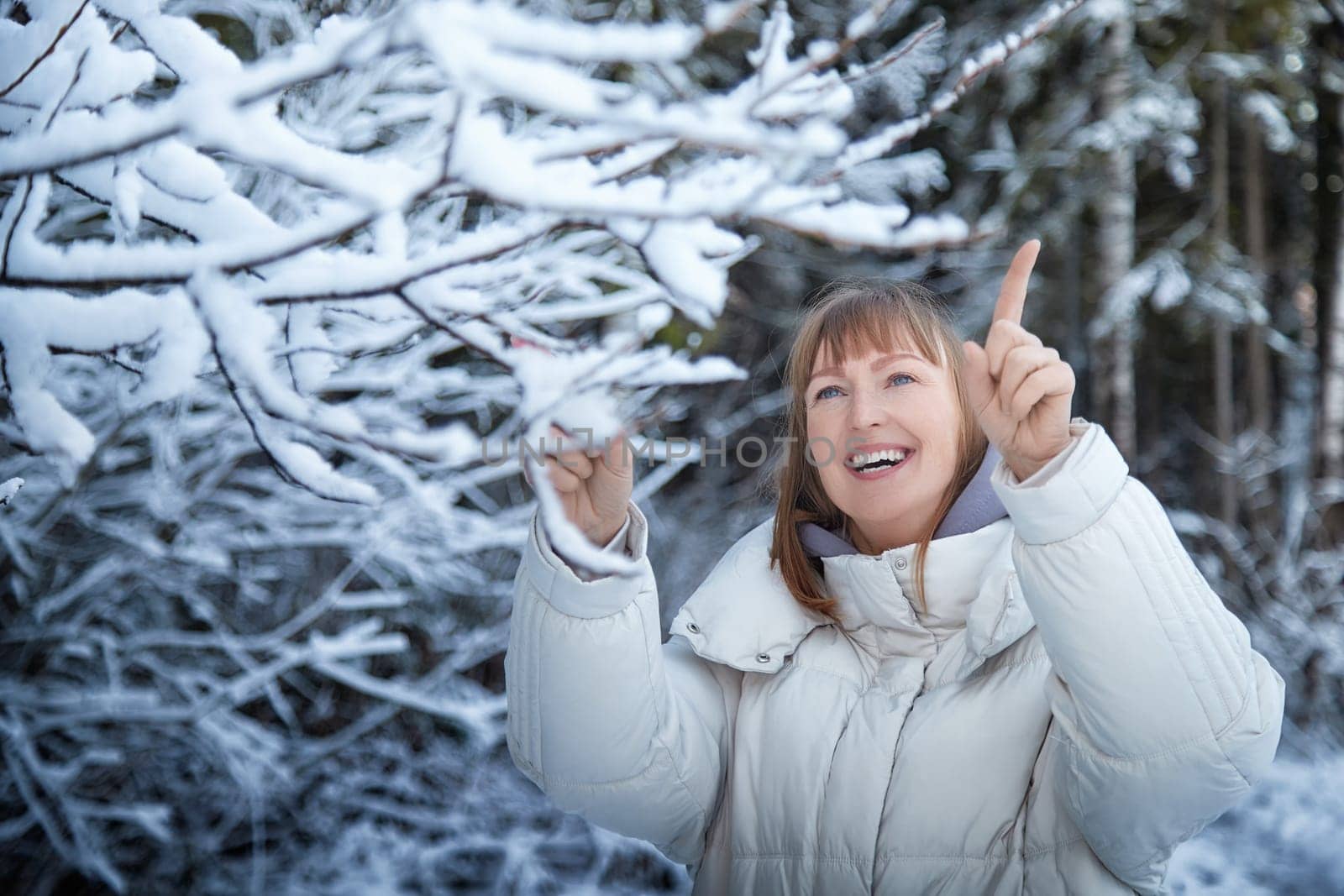 A woman walks through a snow-covered forest, smiling as she points at the snow-laden branches, celebrating the winter season.