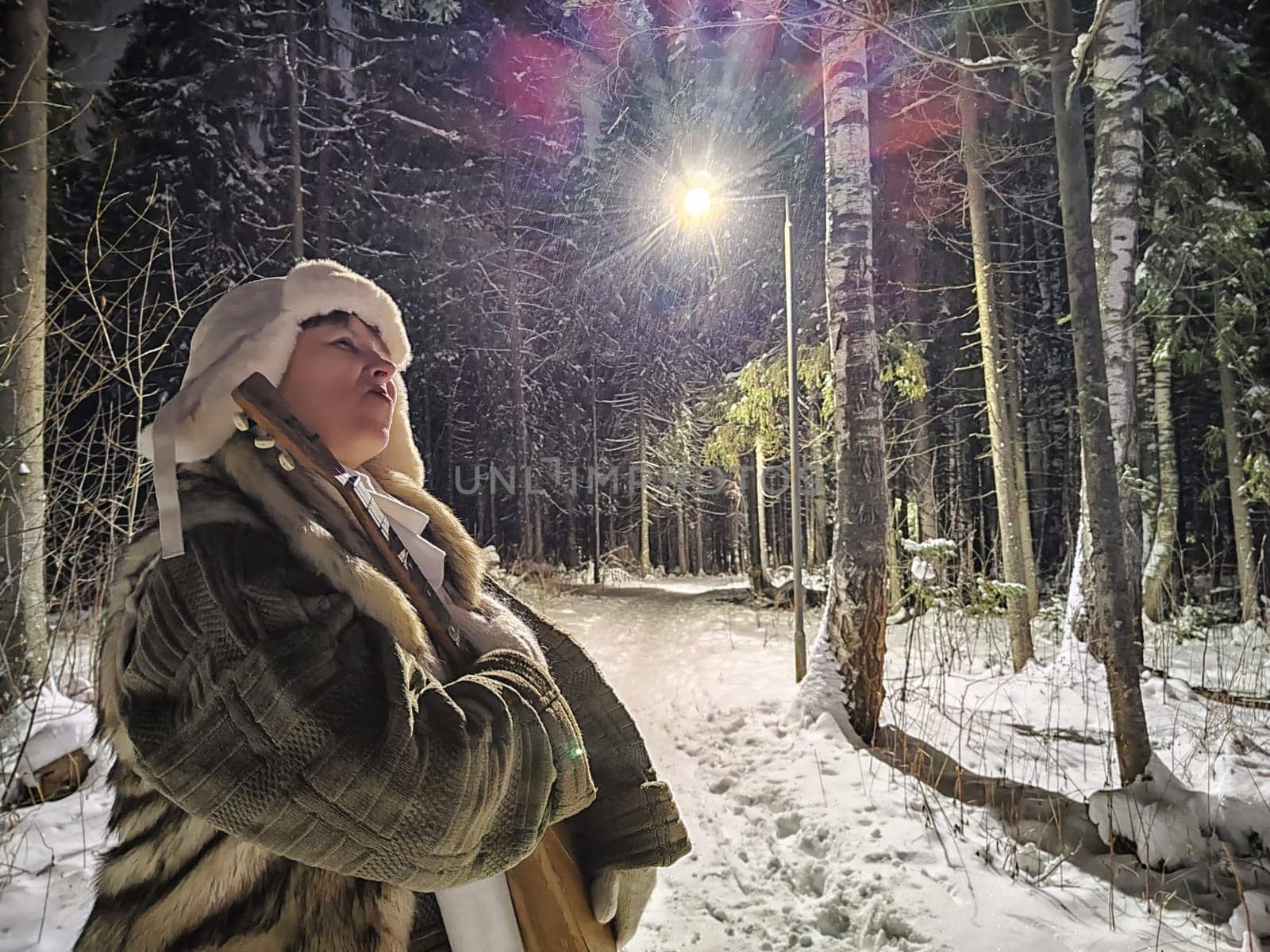 A middle-aged woman strolls through a snowy forest at night, wearing a funny hat and enjoying an unprofessional modeling shoot.