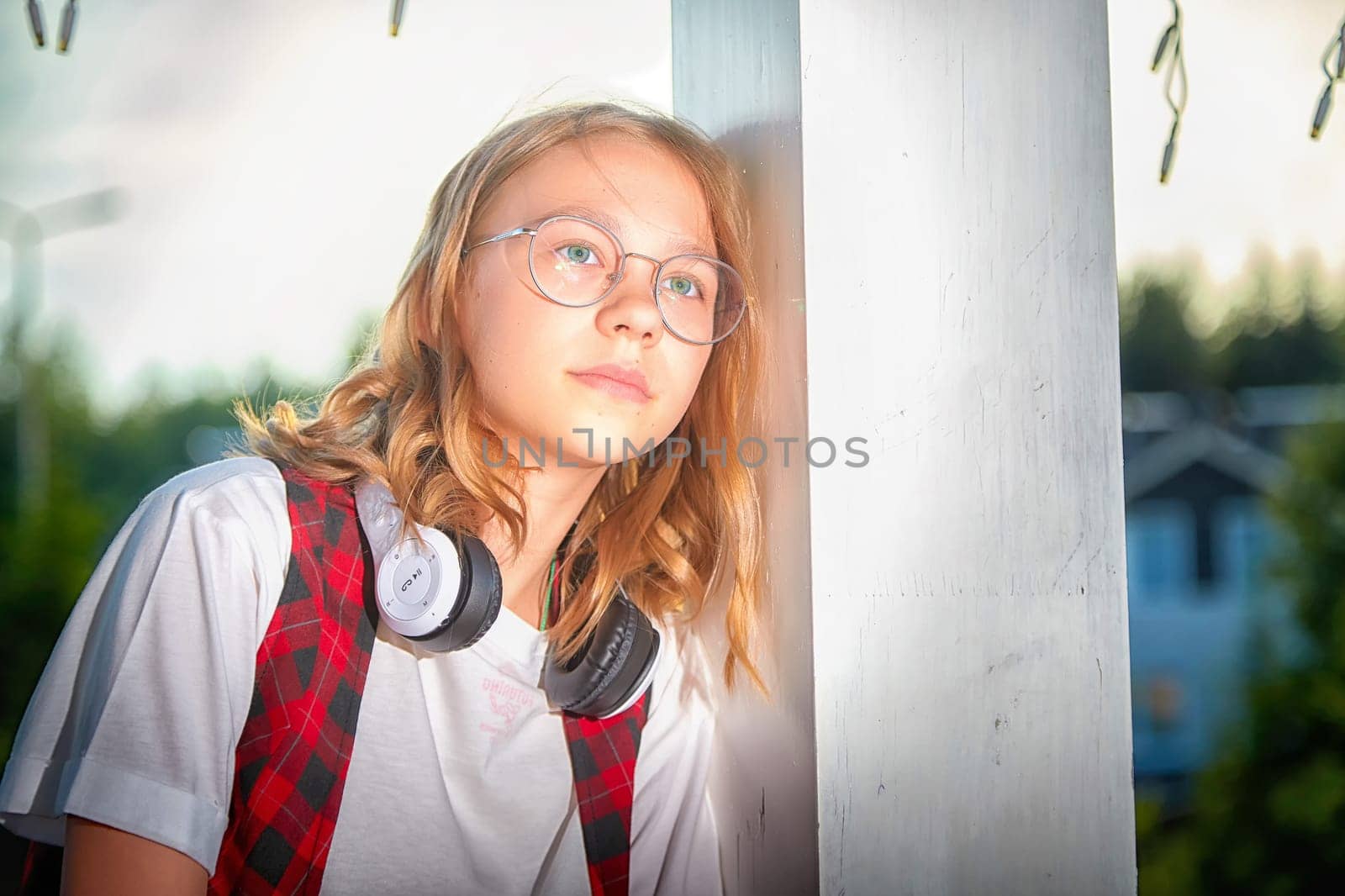 A young individual with glasses rests against a white post, wearing headphones and gazing into the distance during daytime.