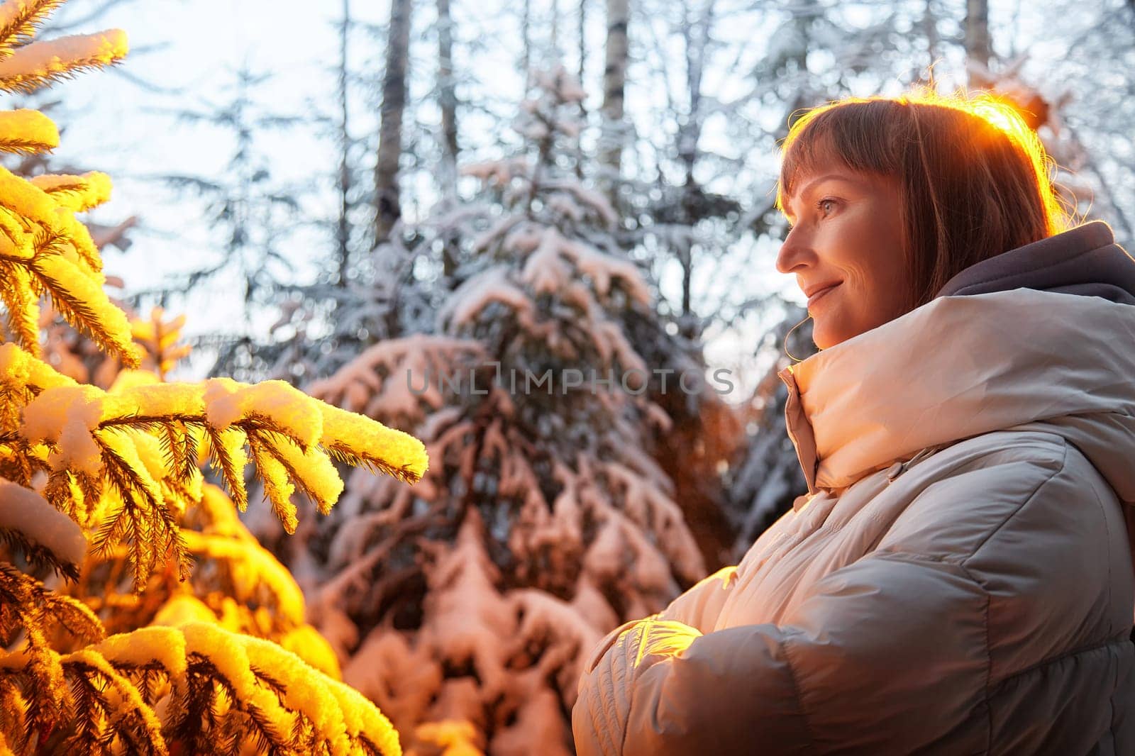 Middle-aged woman enjoys a peaceful walk in a snowy forest while surrounded by shimmering Christmas trees and winter beauty.