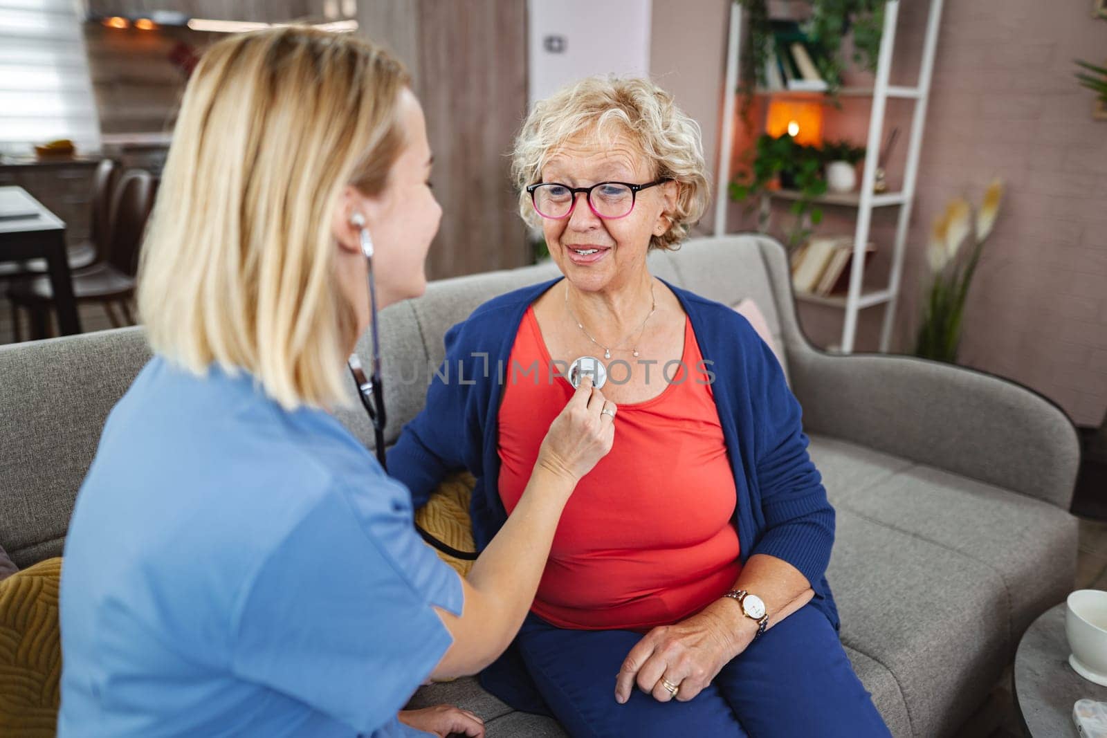 A nurse attentively checks the heartbeat of a smiling senior woman indoors.