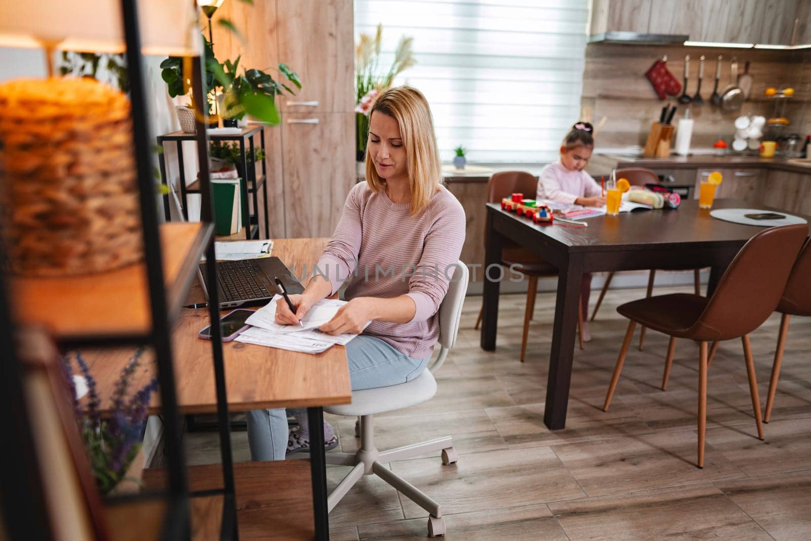 A mother writes at her desk while her daughter crafts nearby.