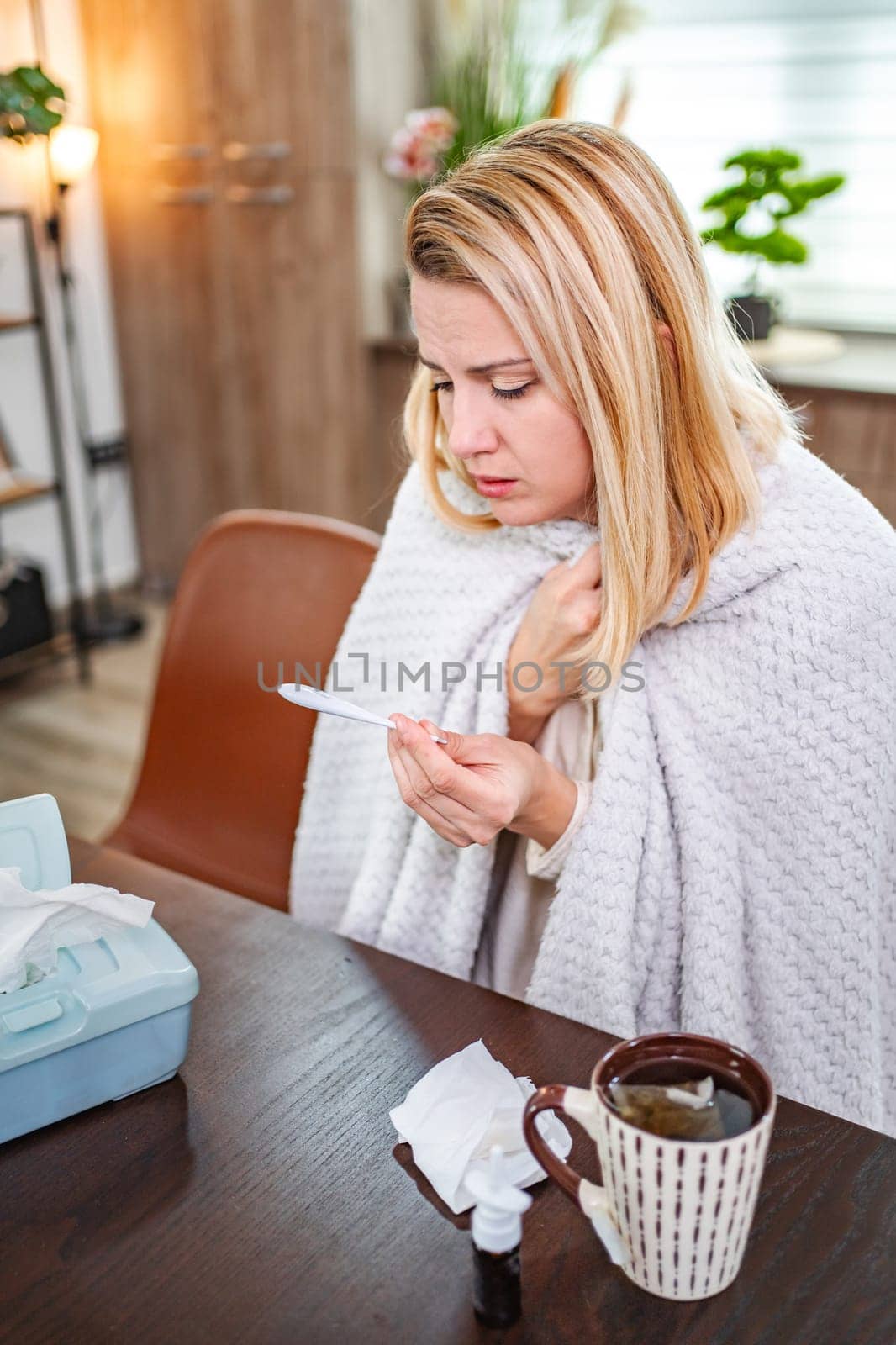 A woman checks her temperature at a table, wrapped in a blanket.