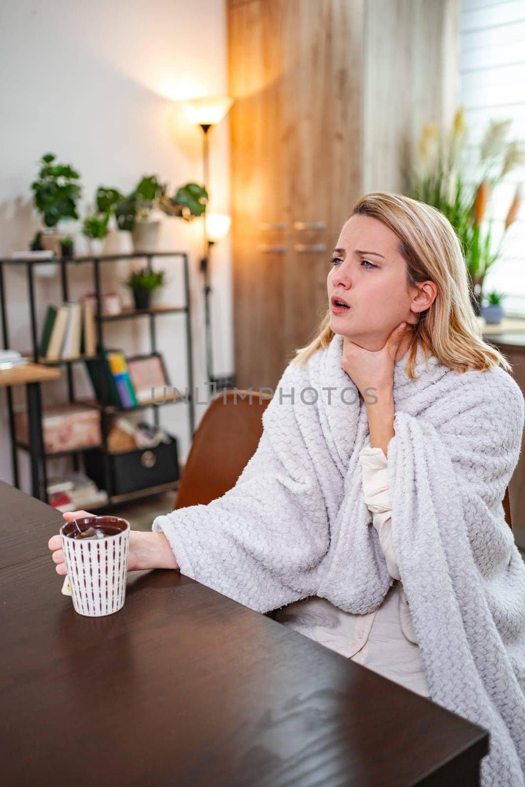 A woman sits at a table, wrapped in a soft blanket, with a pained expression.