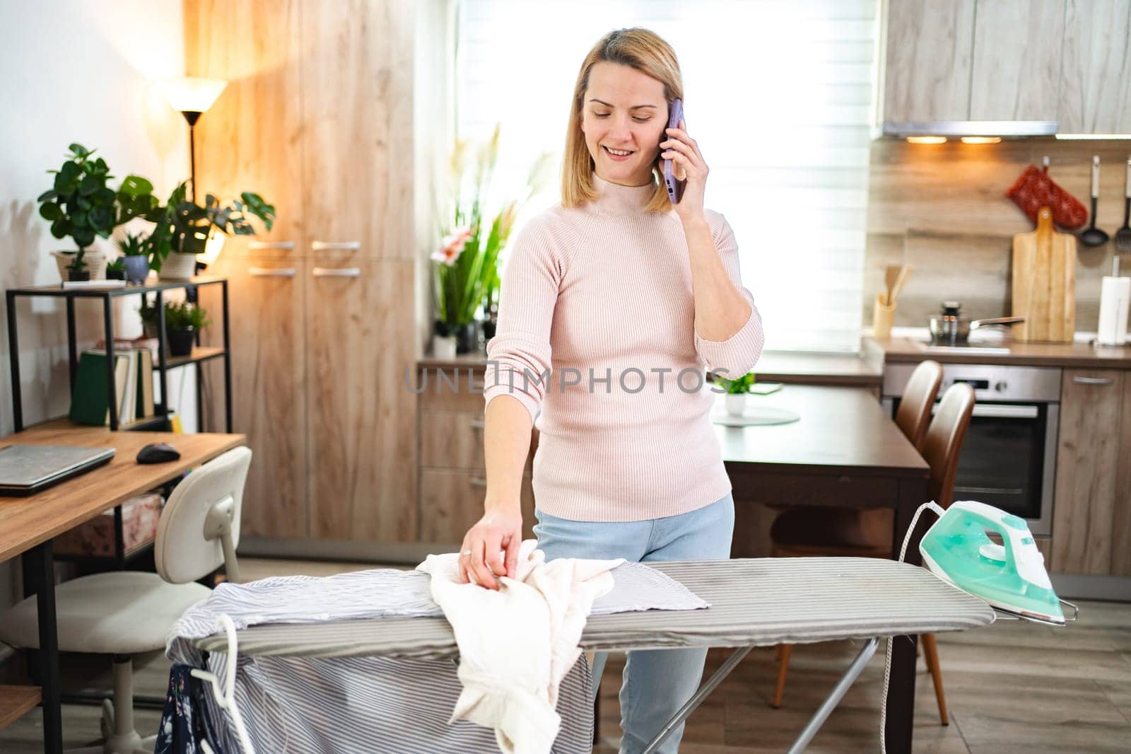 Woman balances phone call while ironing clothes in modern kitchen space
