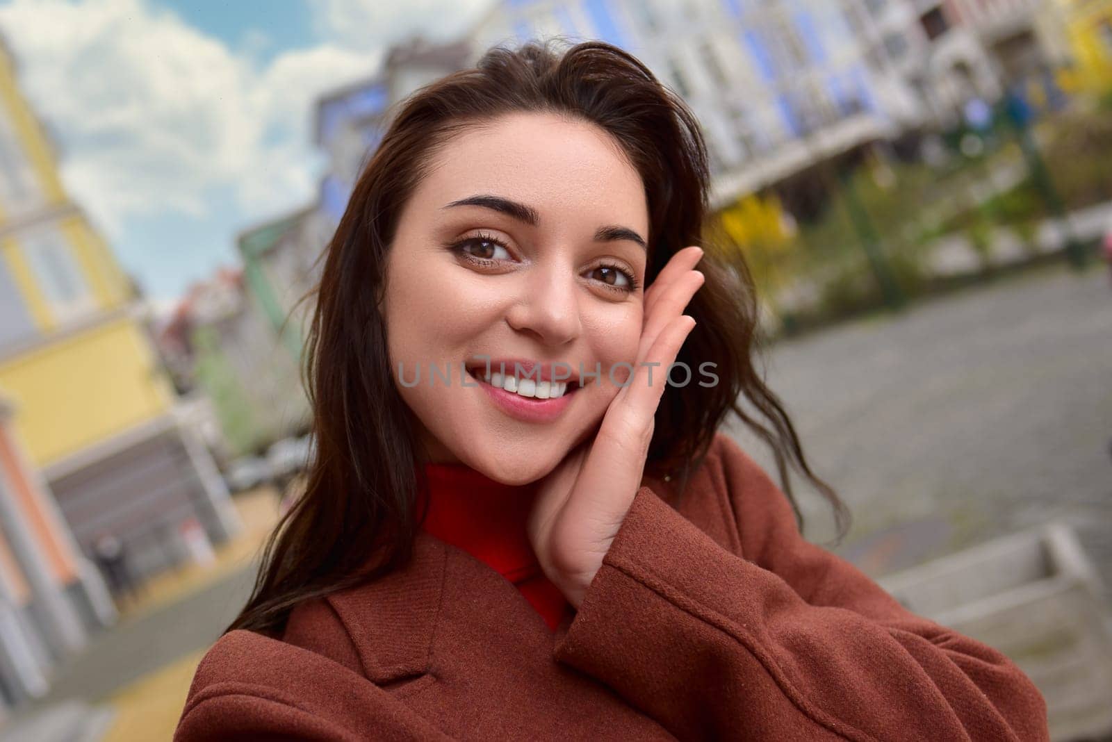 Portrait of a stylish smiling woman in a brown autumn coat takes a selfie on the street.