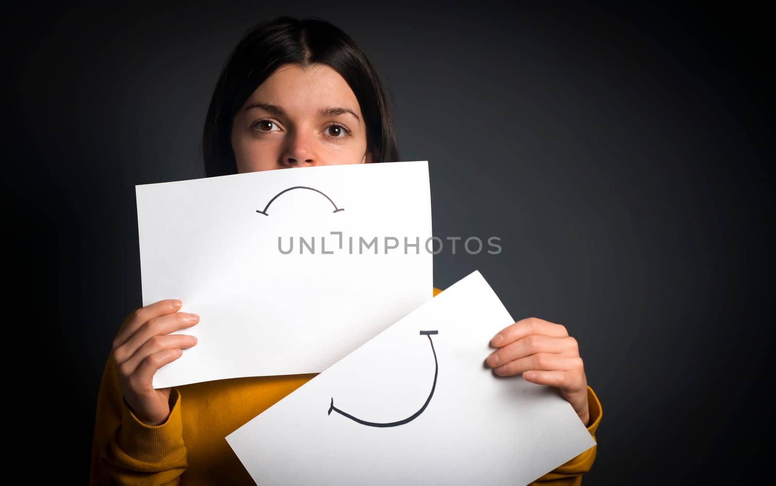 Girl changes, hides emotions, holds 2 papers with sadness and joy, the concept of duality and removal of masks, a woman removes her smiley mask and shows her difficulties with depression.