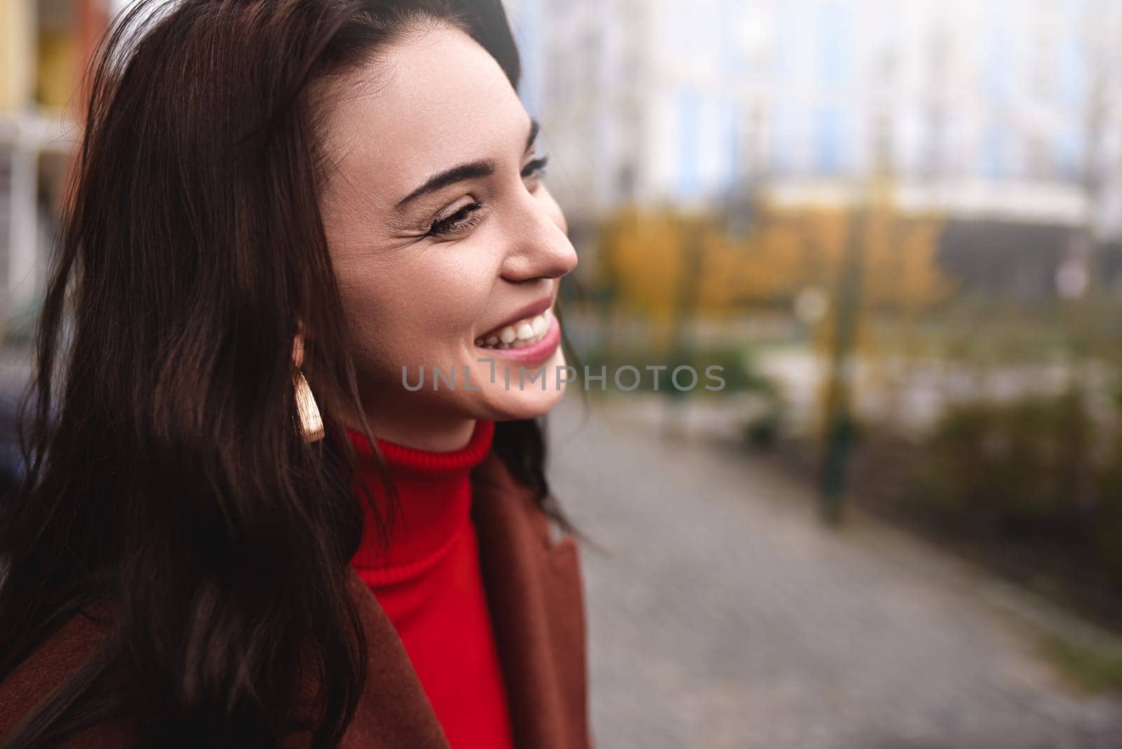 Profile of a young stylish woman in a warm autumn coat, smiling sincerely while walking down the street.