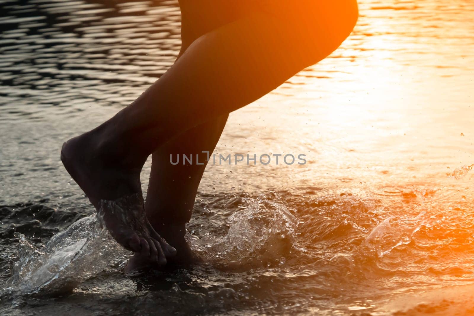 A person runs along the beach on the water, the sea or ocean in the rays of the sun, an athlete man spends time happily, is training, jogging outdoors, close-up view of the legs and splashes.