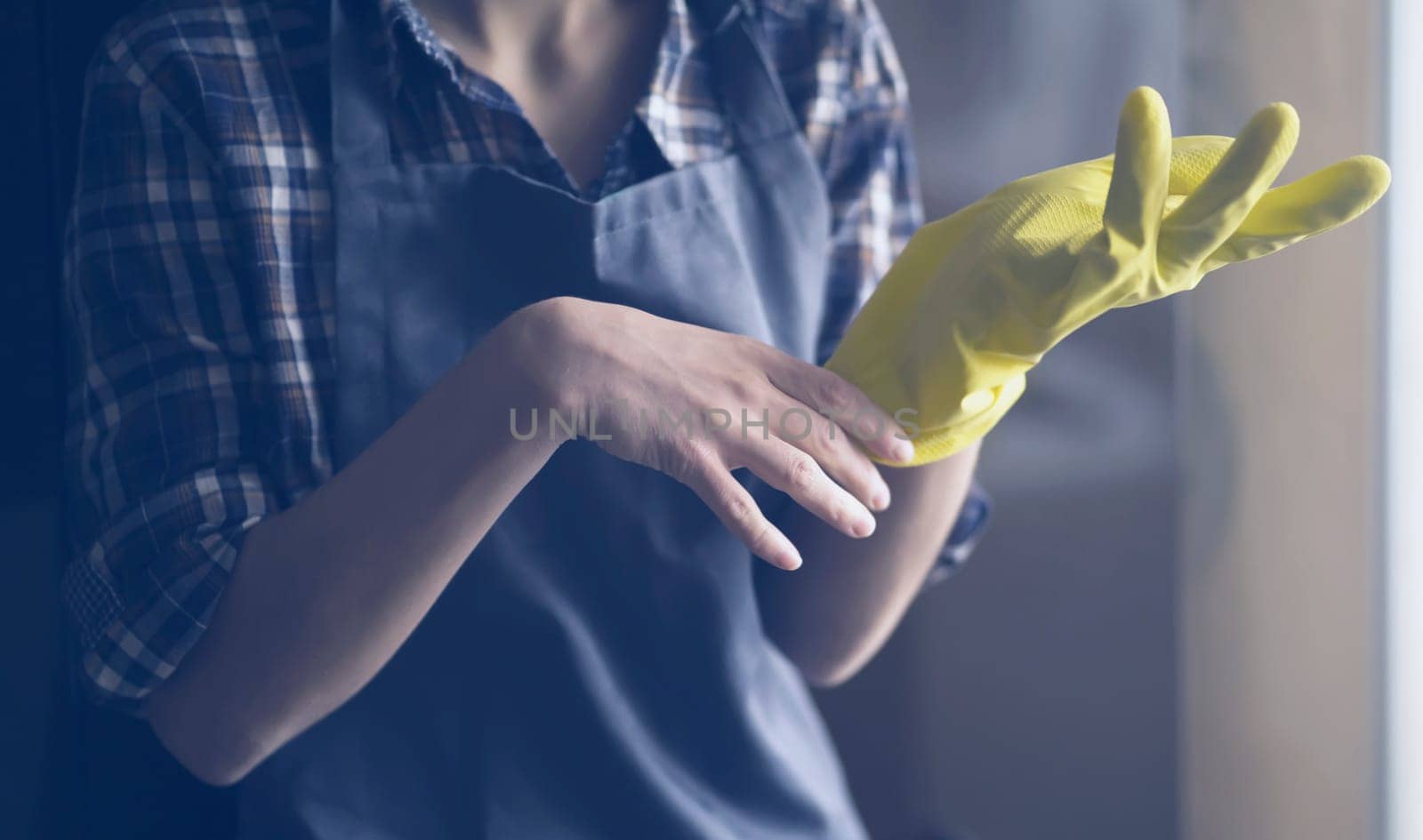 A young girl in a plaid shirt and apron puts yellow rubber gloves on her hands to start cleaning her house and create comfort. Housekeeper with gloves doing disinfection.