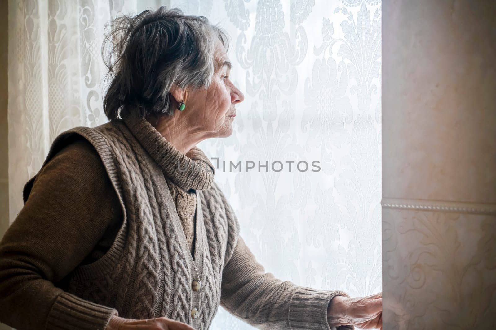 An elderly gray-haired woman in a casual knitted sweater looks out the window of her house, an old grandmother is waiting for guests, stands against the background of a window with lace curtains.