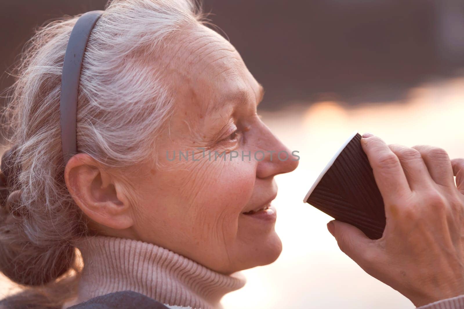 A beautiful elderly woman with gray hair and wrinkles on her face smiles and enjoys coffee or tea against the background of the river at sunrise or sunset in the city during a walk, portrait closeup.