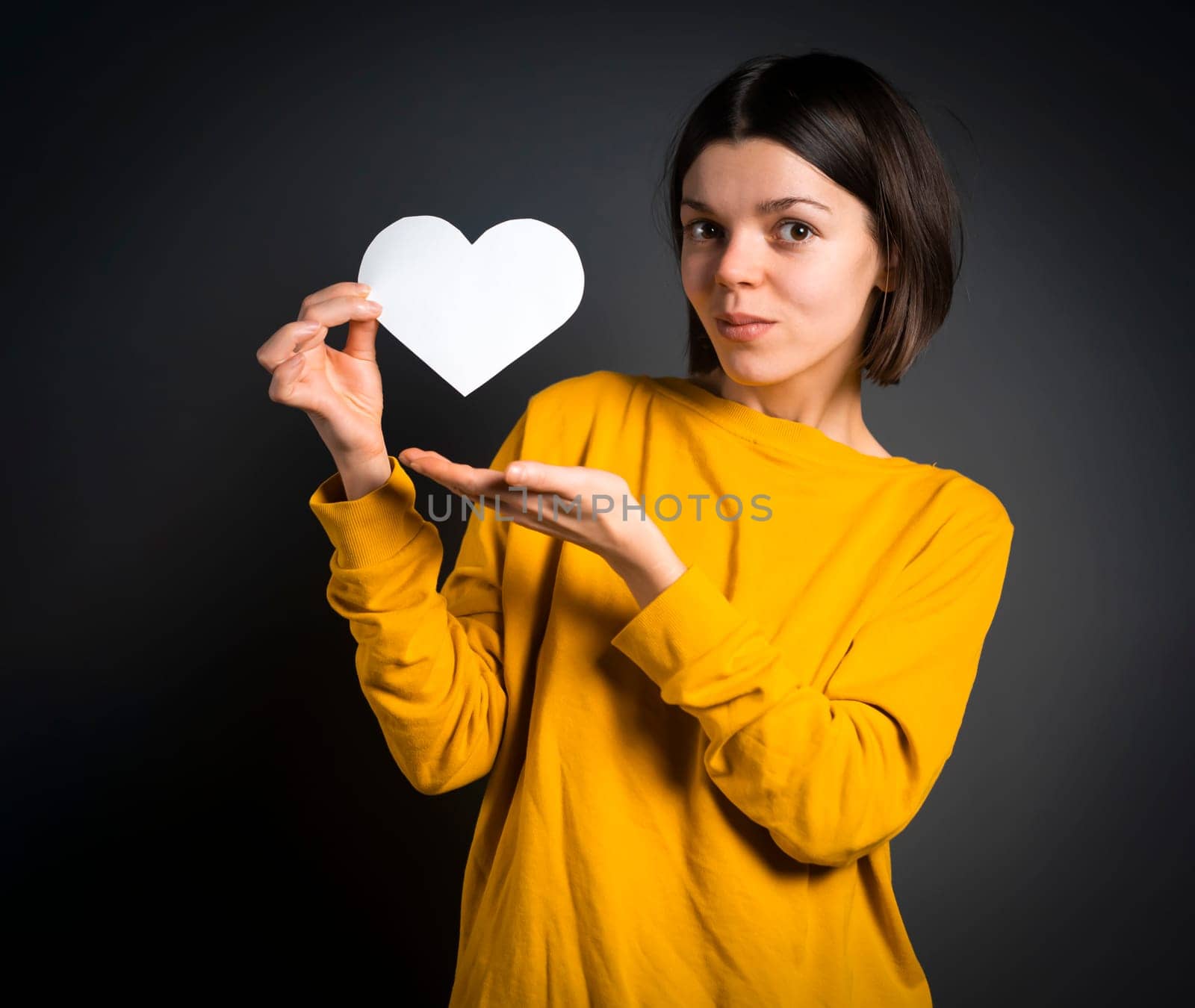 A young girl holds a white paper heart - a postcard or shows feelings of love. A woman with a joyful surprised face before the holiday and in search of love, relationships or shares her impressions.