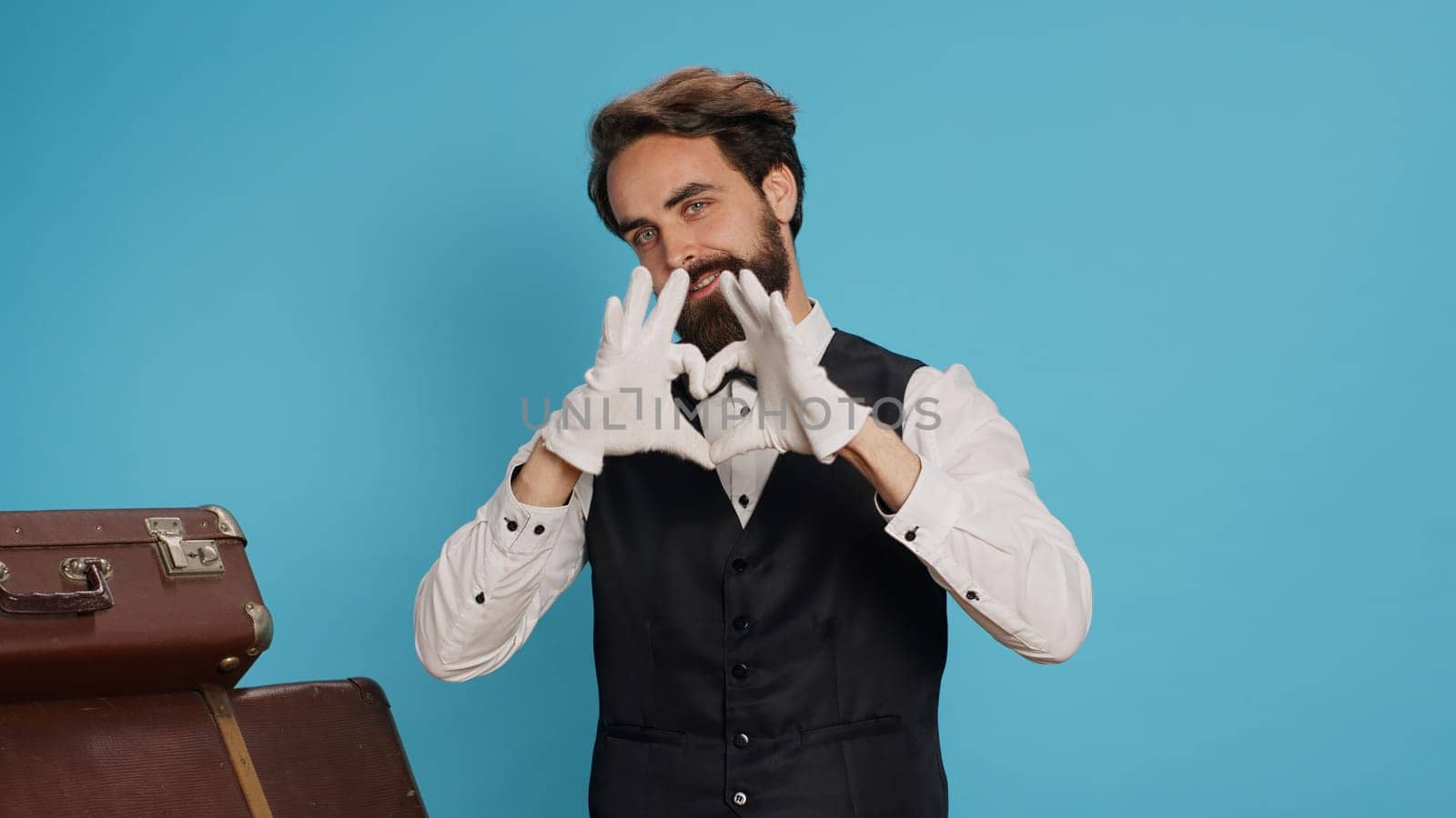 Cheerful bellhop does heart shaped sign in studio, expressing romantic honest feelings and flirting while he wears formal suit. Lovely employee showing romance symbol for love, sincere emotions.