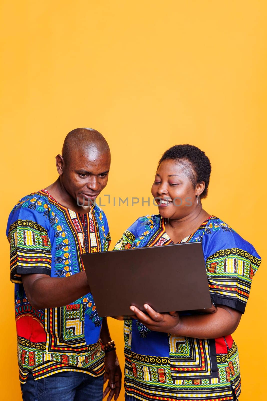 Smiling black man and woman sharing one laptop for work and online pastime activity. Cheerful african american couple wearing ethnic clothes using portable computer in studio