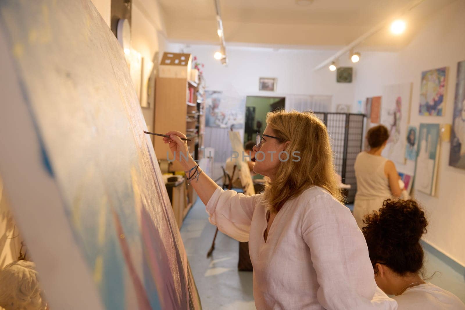 Side portrait of a Caucasian creative mature blonde woman working on painting at her art studio. Art class. Education. Creative occupation