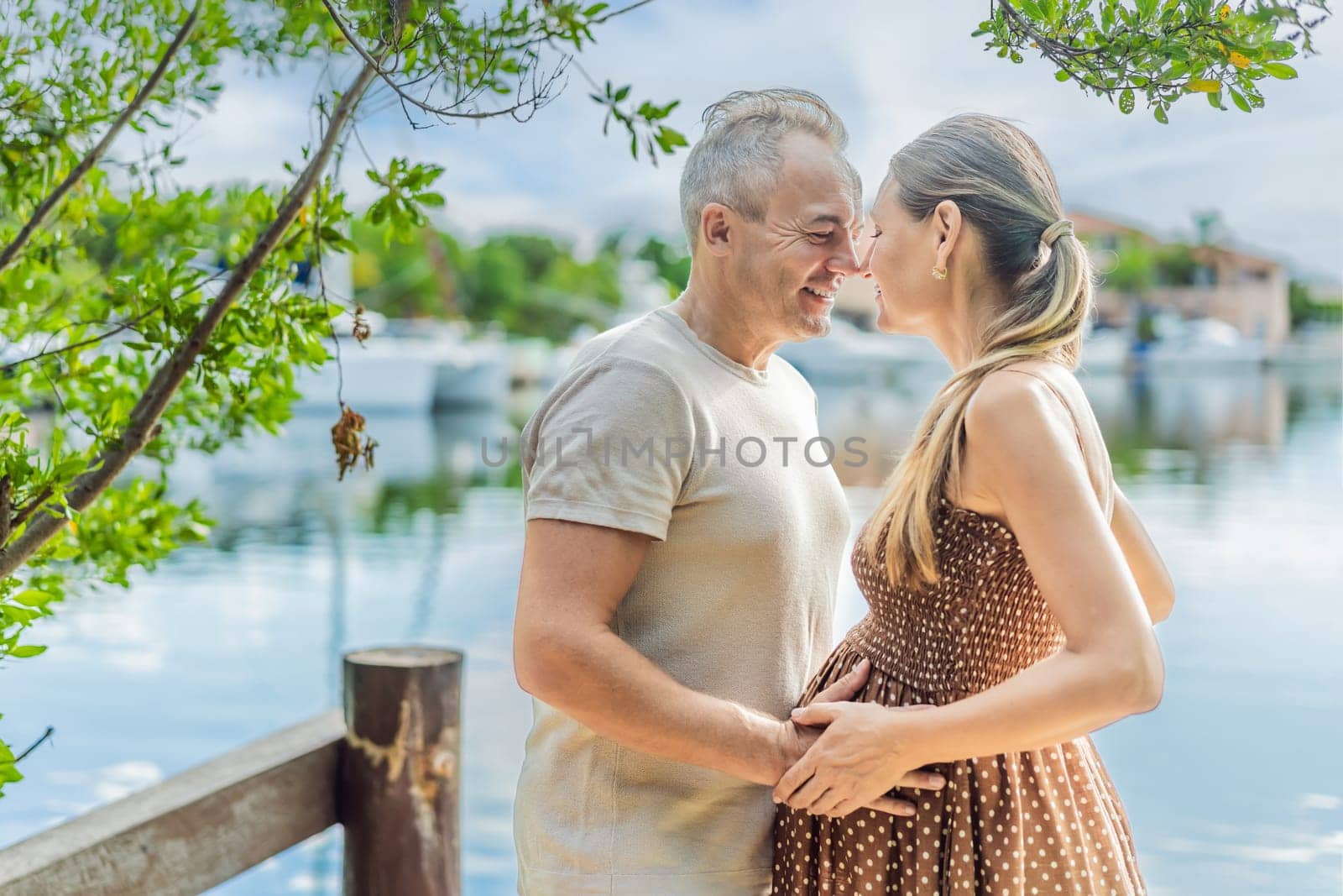 A happy, mature couple over 40, enjoying a leisurely walk on the waterfront, their joy evident as they embrace the journey of pregnancy later in life by galitskaya